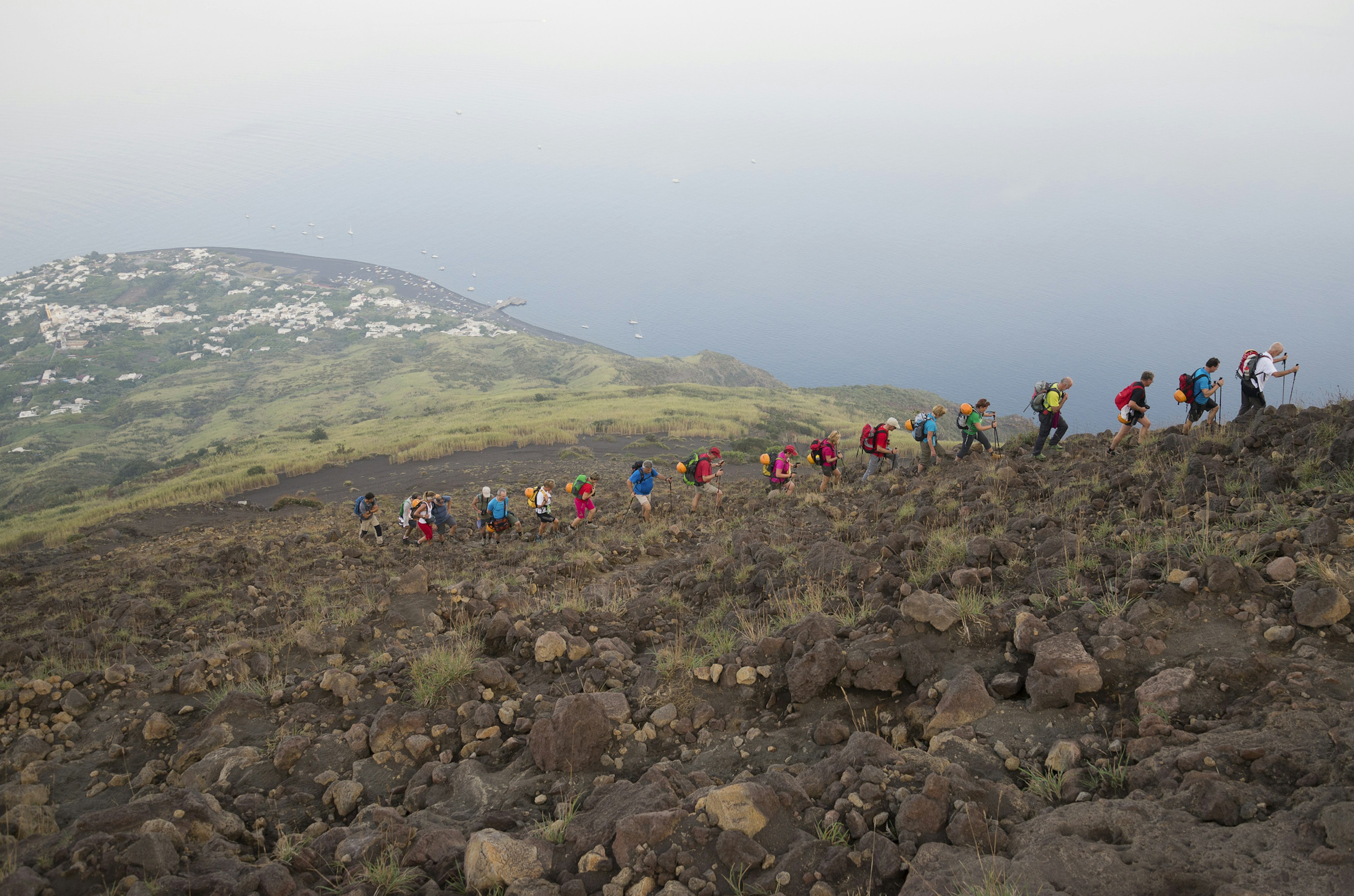 Several hikers in a line following a path up the side of a steep volcano. The town and coastline are in the distance below them