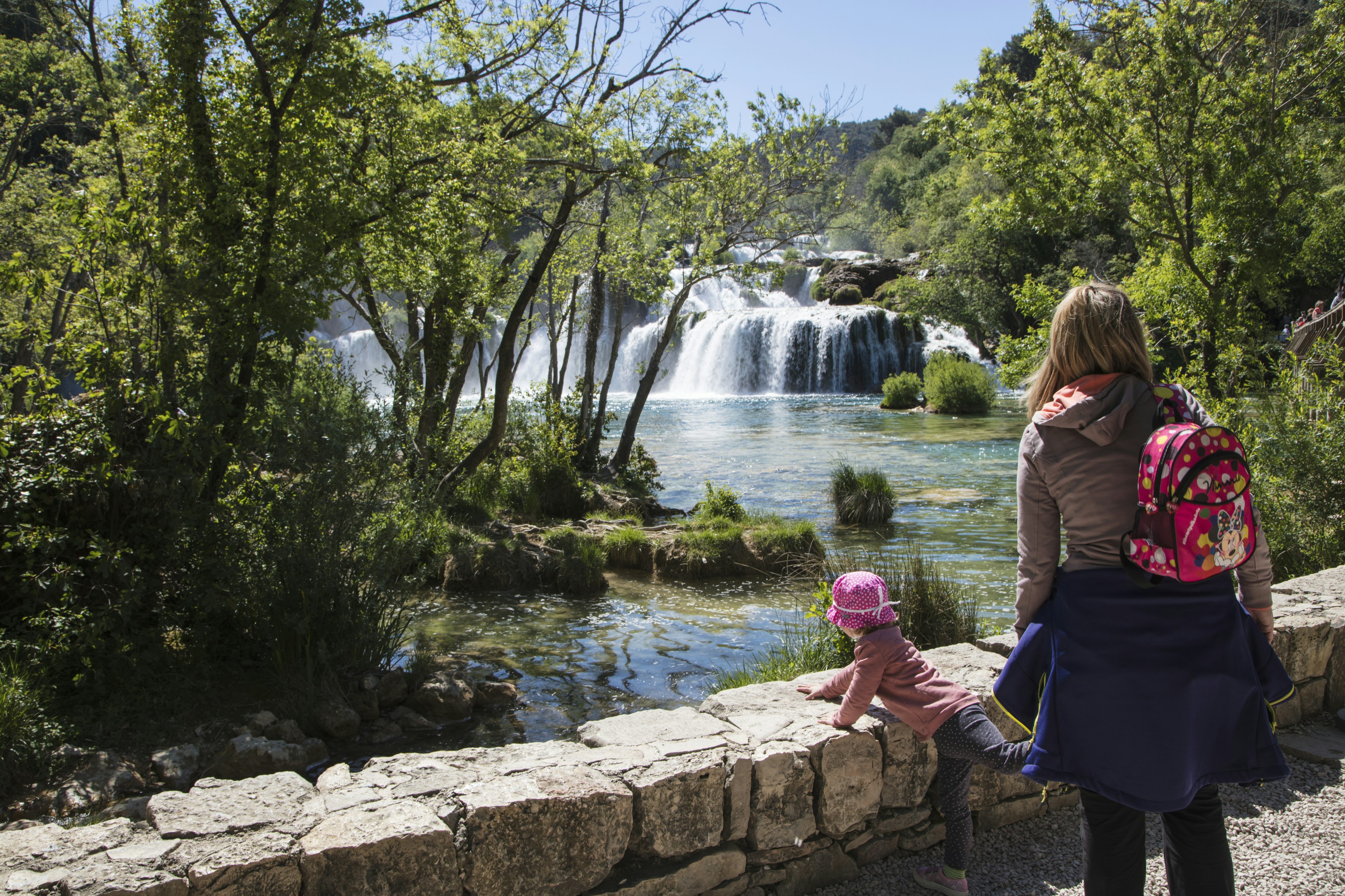 A mother and small child overlook waterfalls plunging into a wide turquoise pool