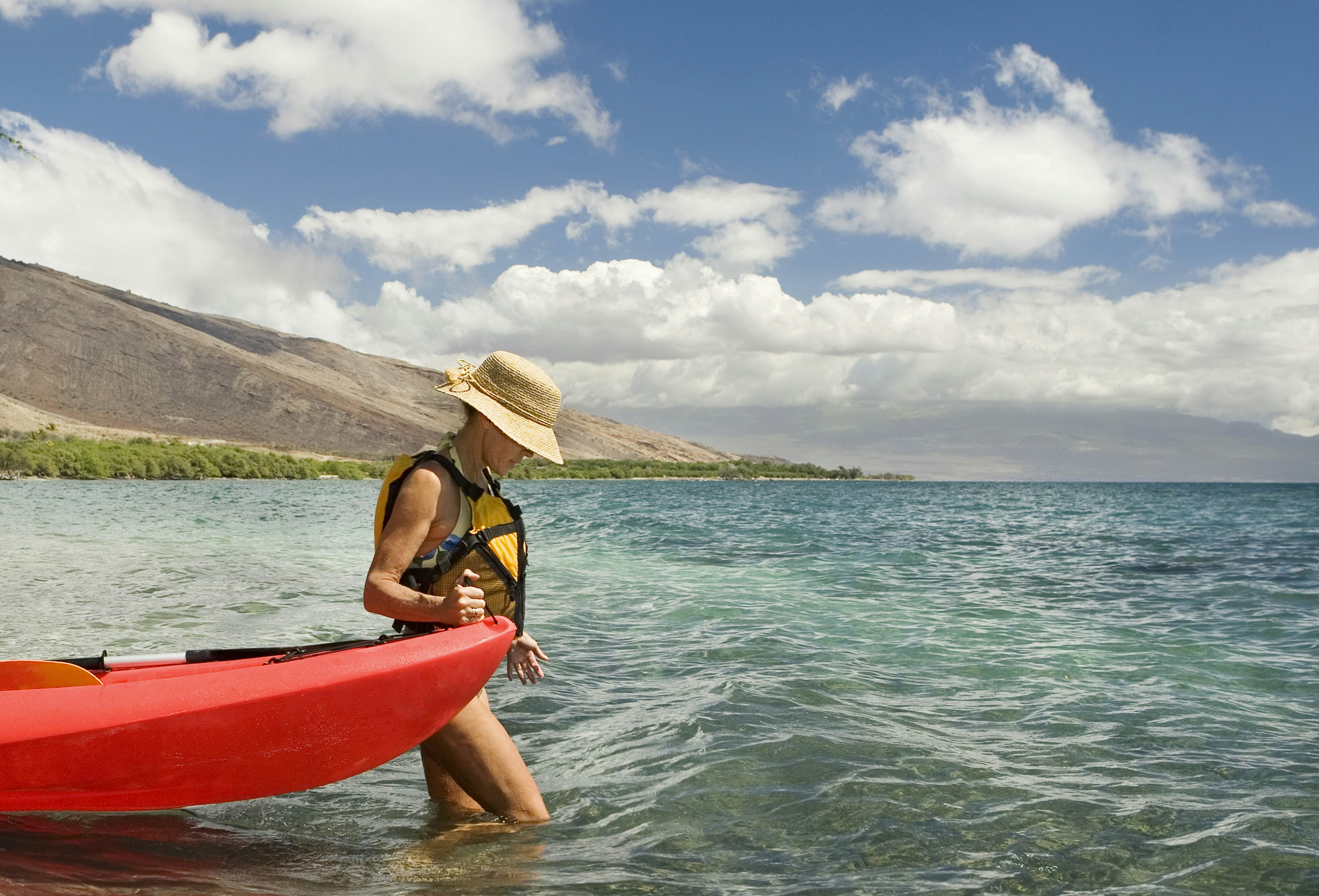 A woman wearing a hat and life vest guides a red canoe into the water in Maui