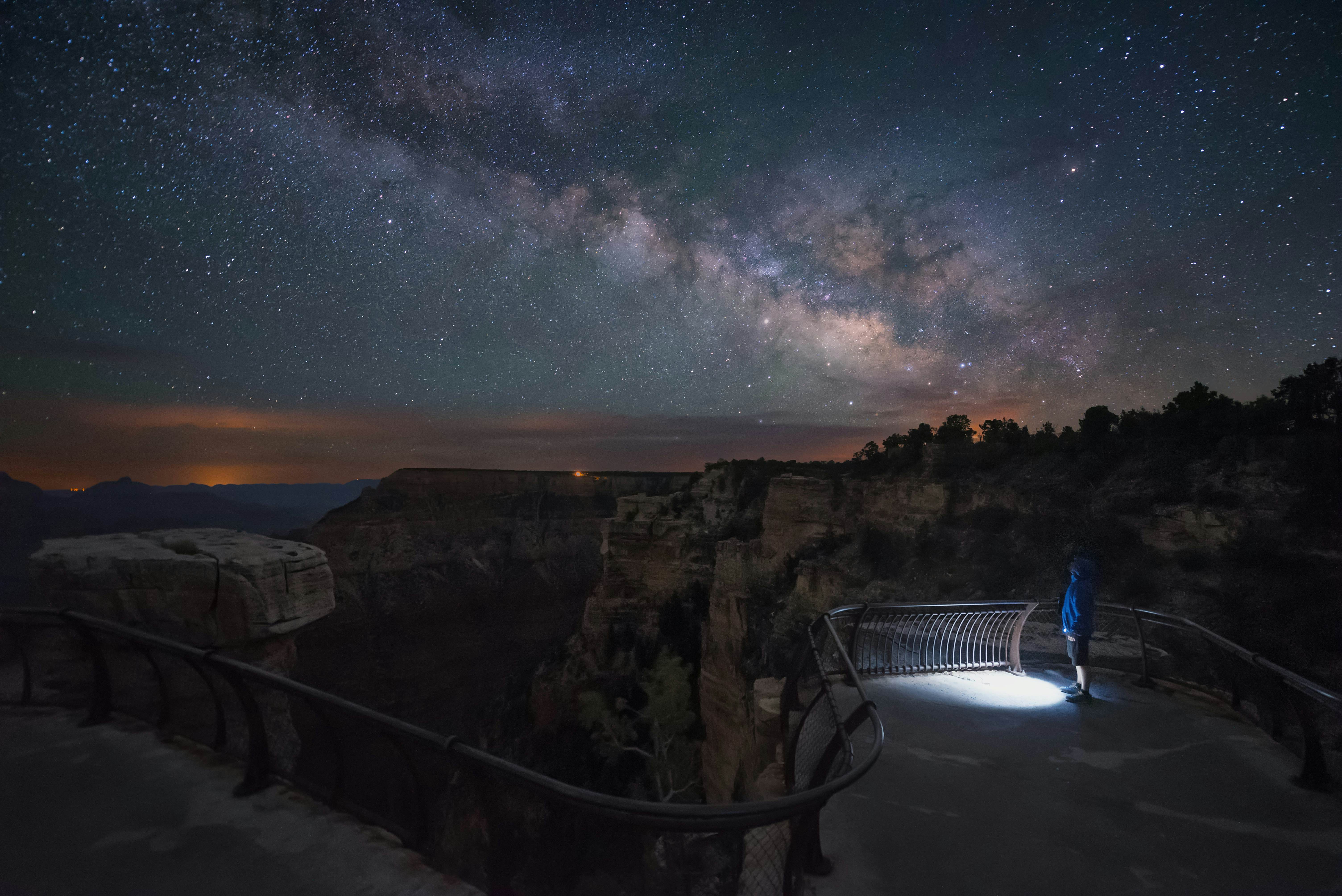 A solo figure with a torch stands at a lookout point on the edge of a canyon at night with a starry sky overhead