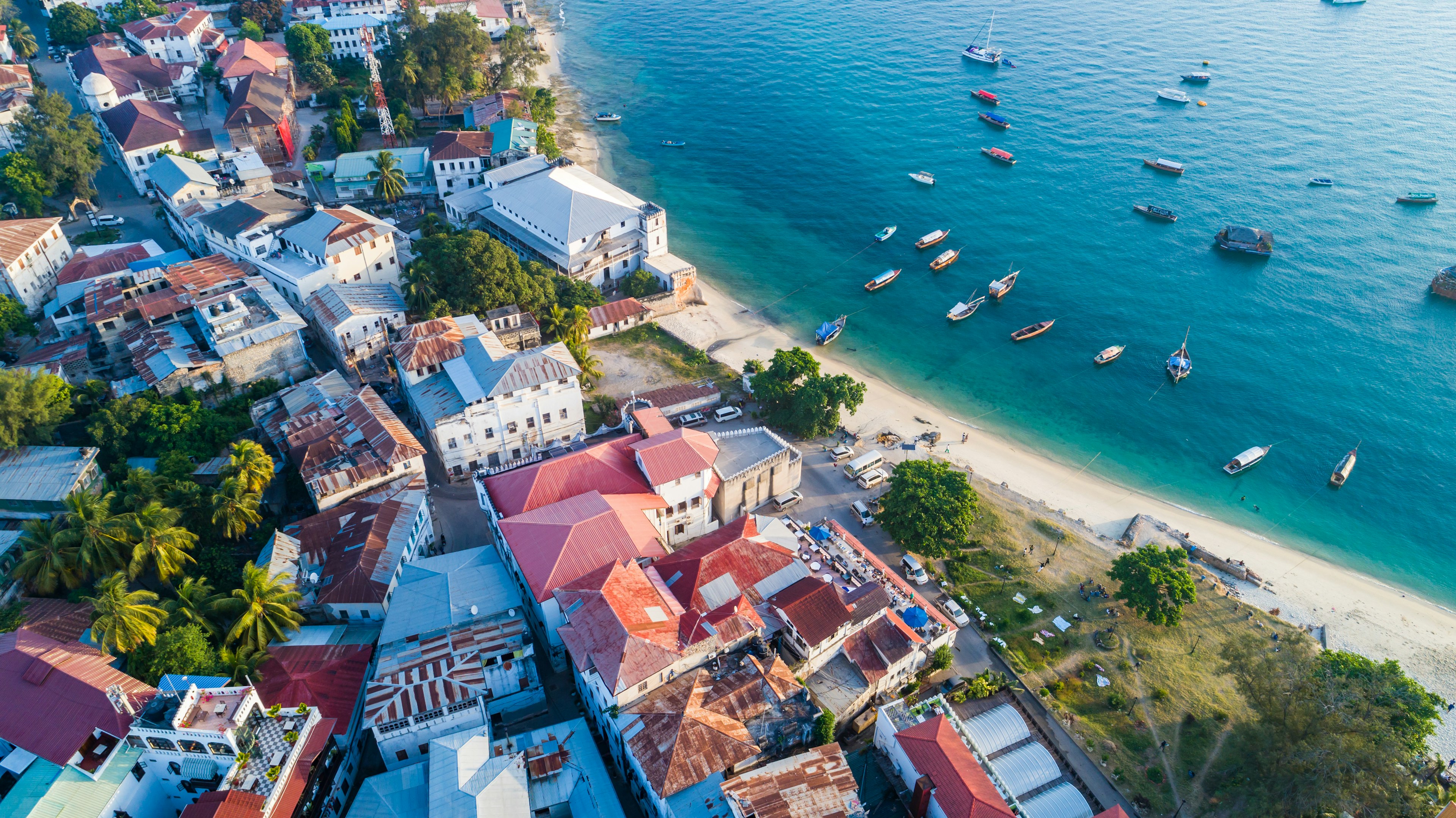 An ariel view over Stone Town and its adjacent beach, with boats floating in blue waters; the town is a jumbled collection of buildings with red, pink and grey roofs.