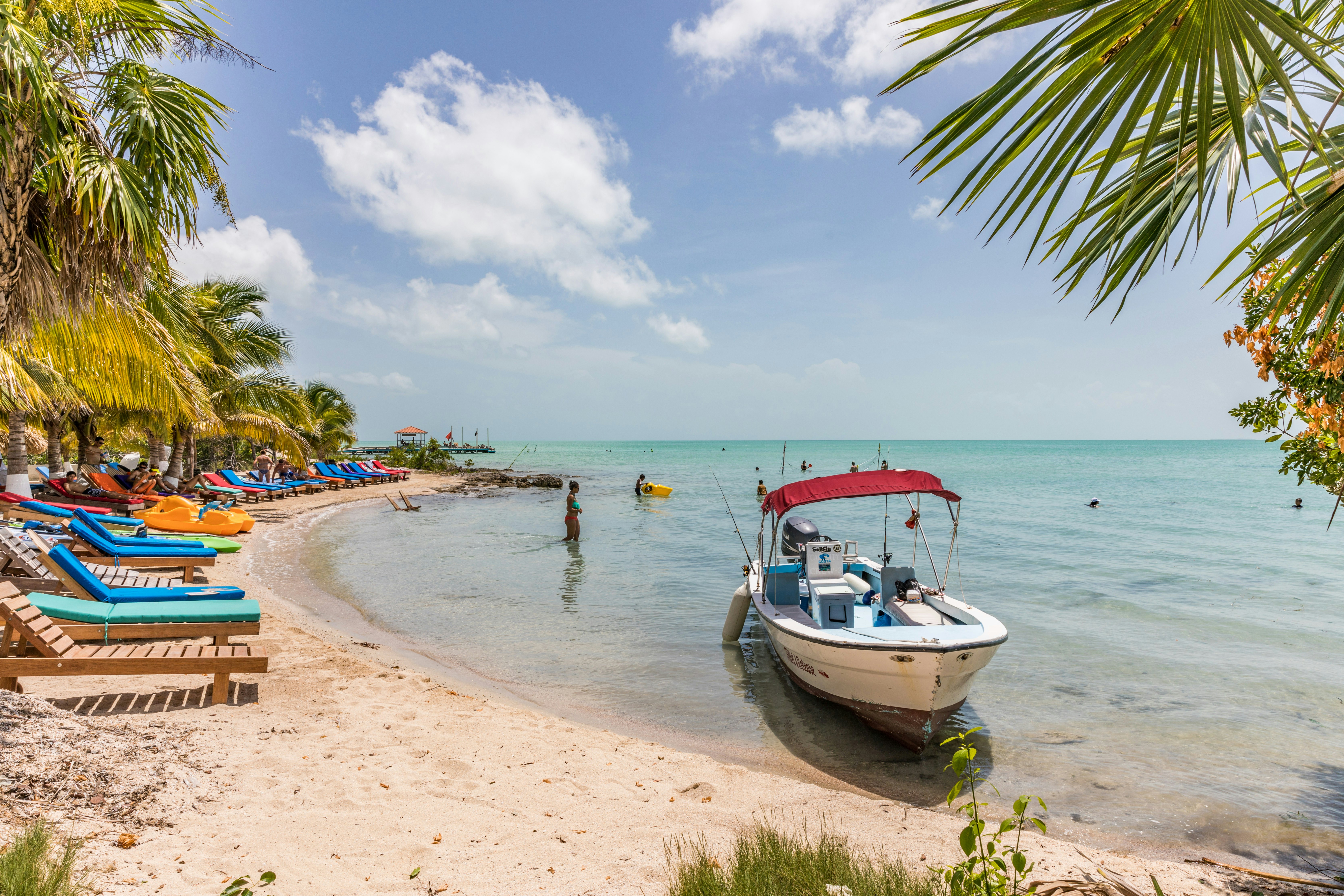 Lounge chairs, a woman in a bikini wading in the water, and a boat anchored just off the beach