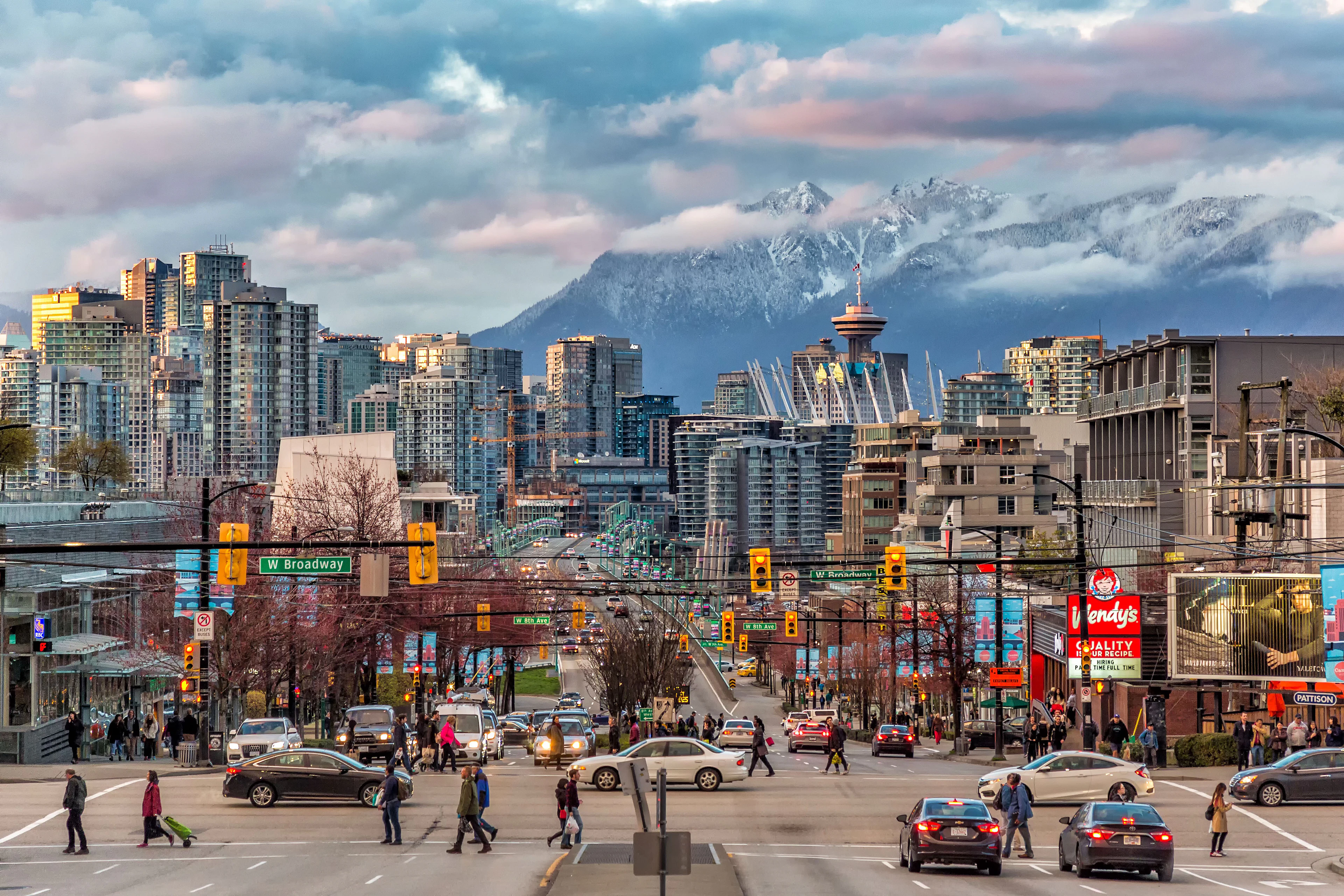 A crossing at Downtown Vancouver where cars wait at the traffic lights and people cross the road during the day with snow-capped mountains across the strait in the background.