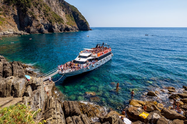 A ferry boat floats in the shallow blue waters off the coast of Riomaggiore in Cinque Terre. The boat is very busy with many people up on deck.