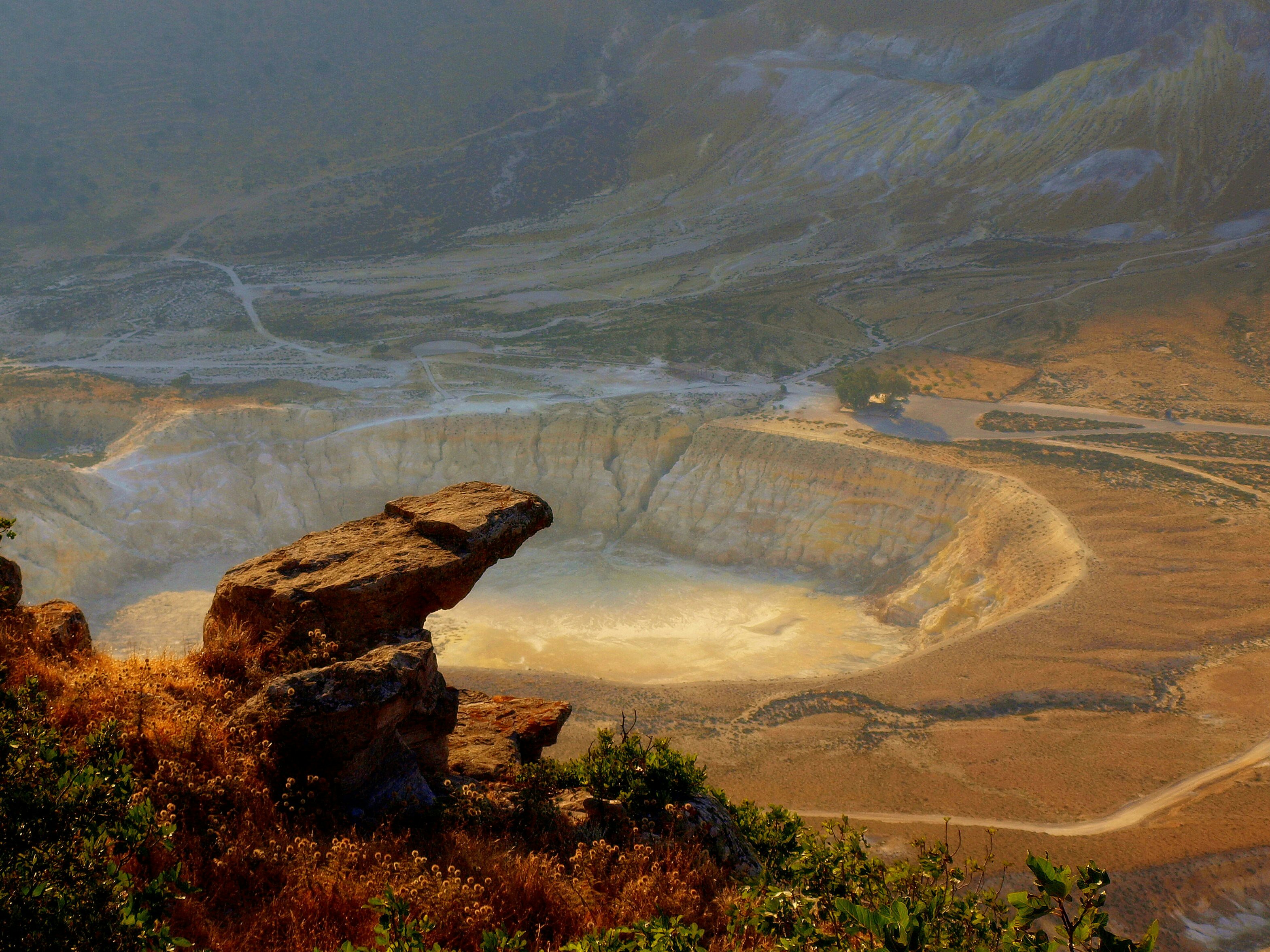 An aerial view of Stefanos Crater, a huge crater in the ground on the island of Nisyros. The exposed white rock gives the crater a lunar-like landscape.