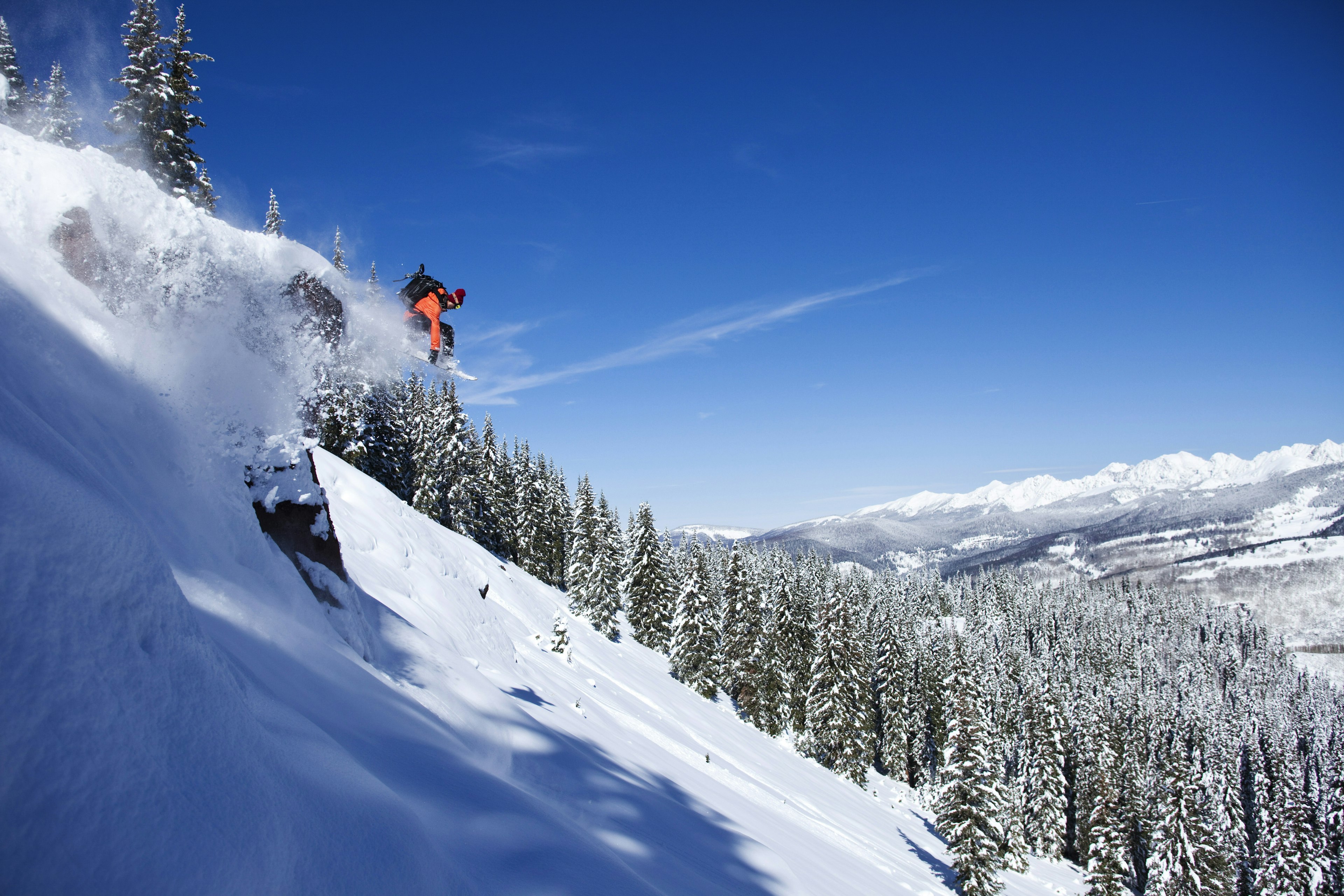 A skier catches air on a steep slope with snow-covered pines and mountains in the distance, Vail