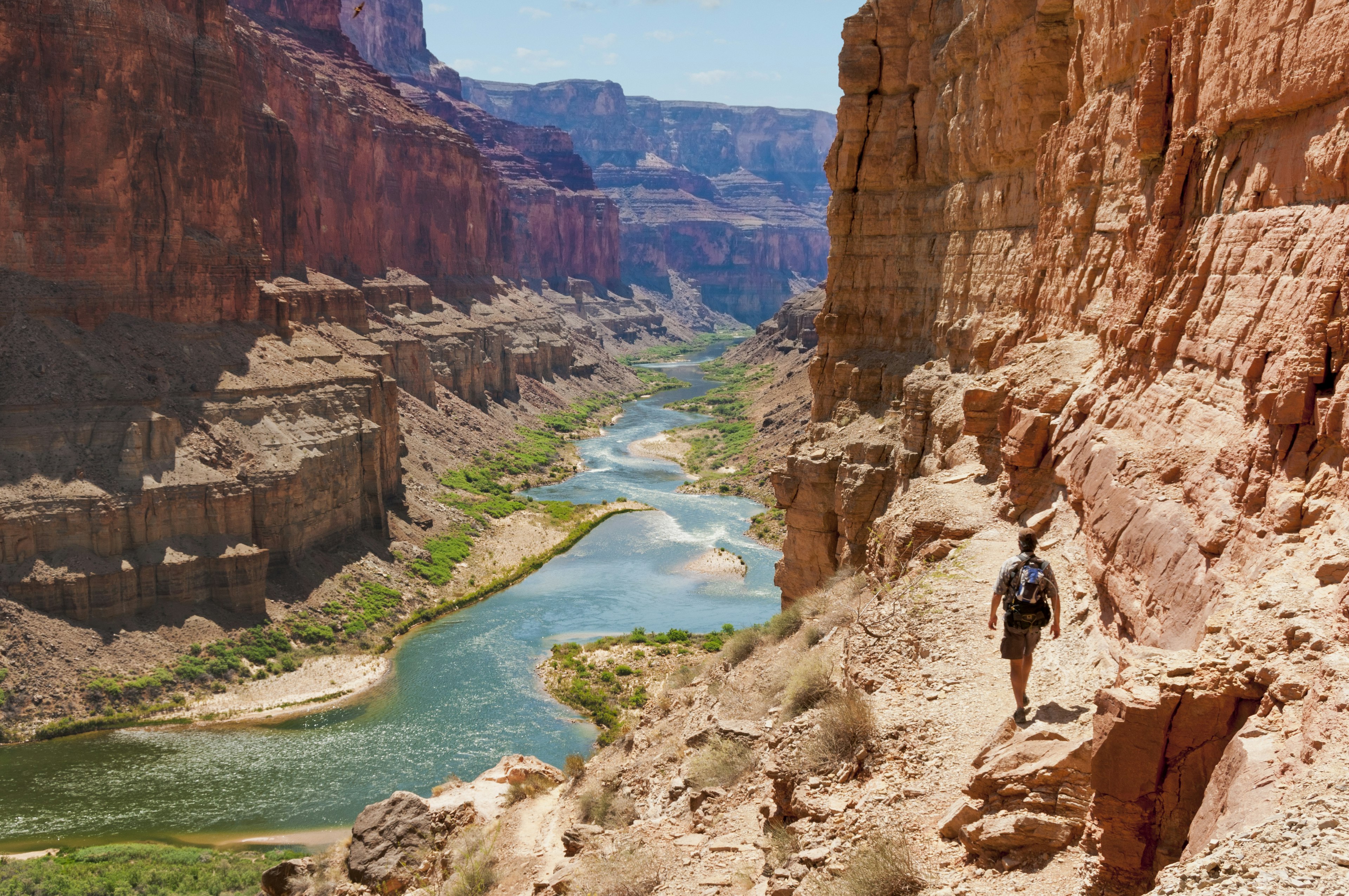 A man hikes a riverside trail through a red-rock canyon