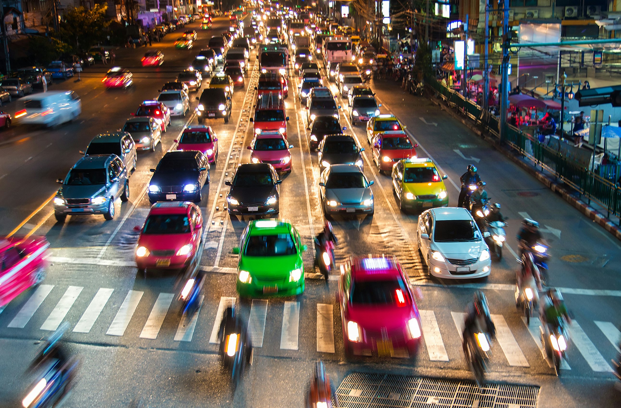 A stream of brightly-colored taxis in green, yellow and pink wait at a junction in Bangkok at night.