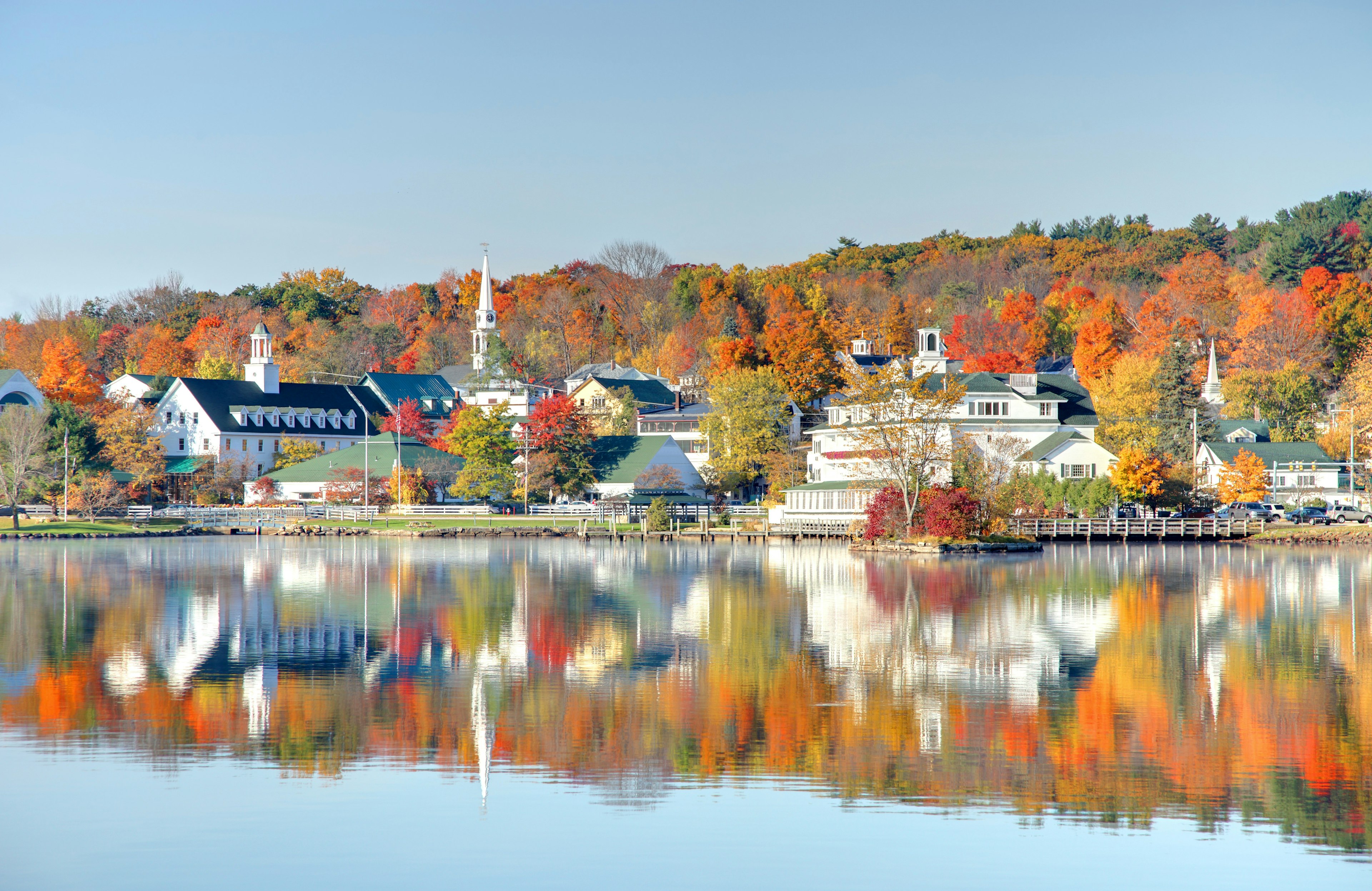 Autumn colors of the woodland behind white buildings are reflected in still lake waters