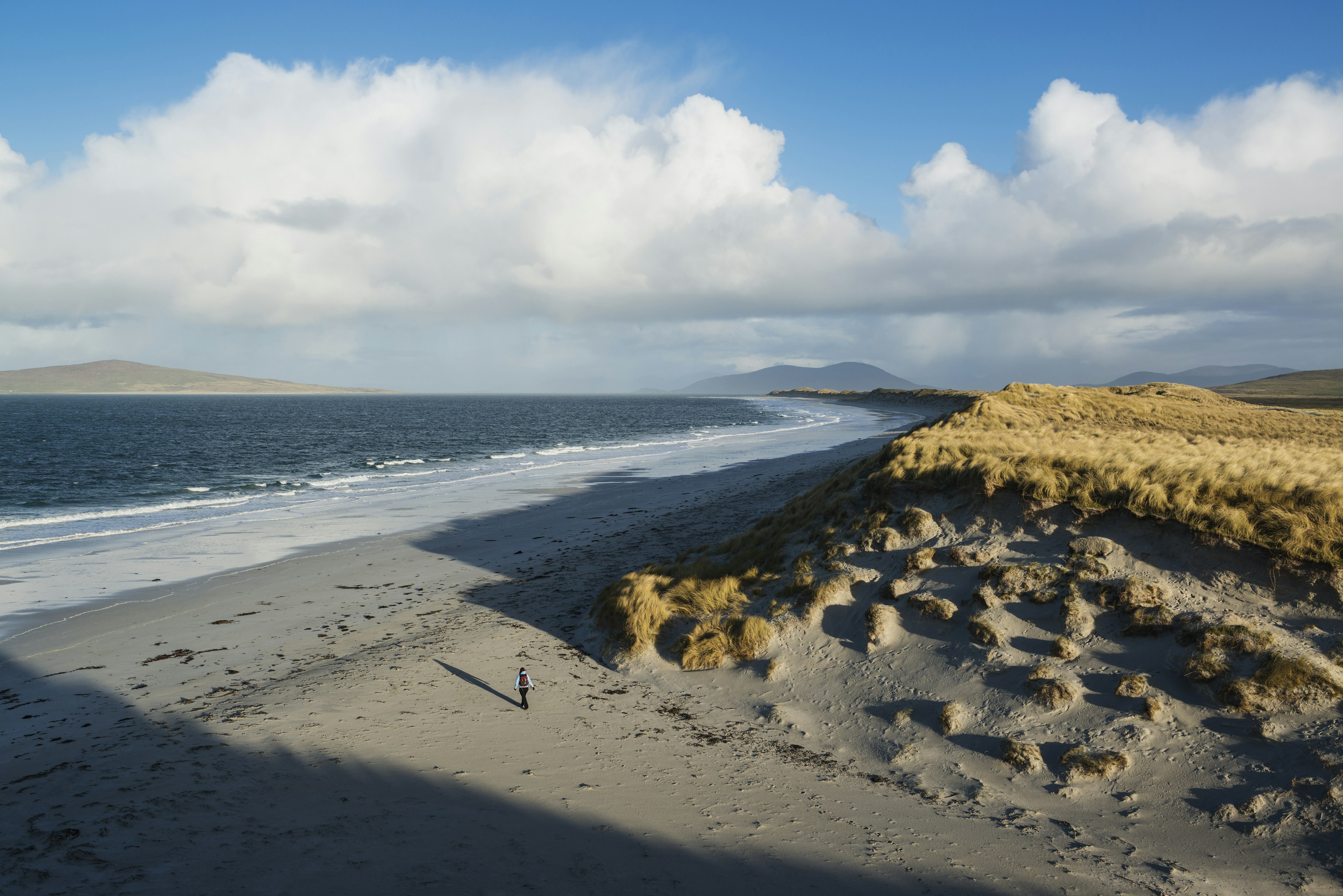 A woman is the sole person on a vast beach backed by grassy dunes