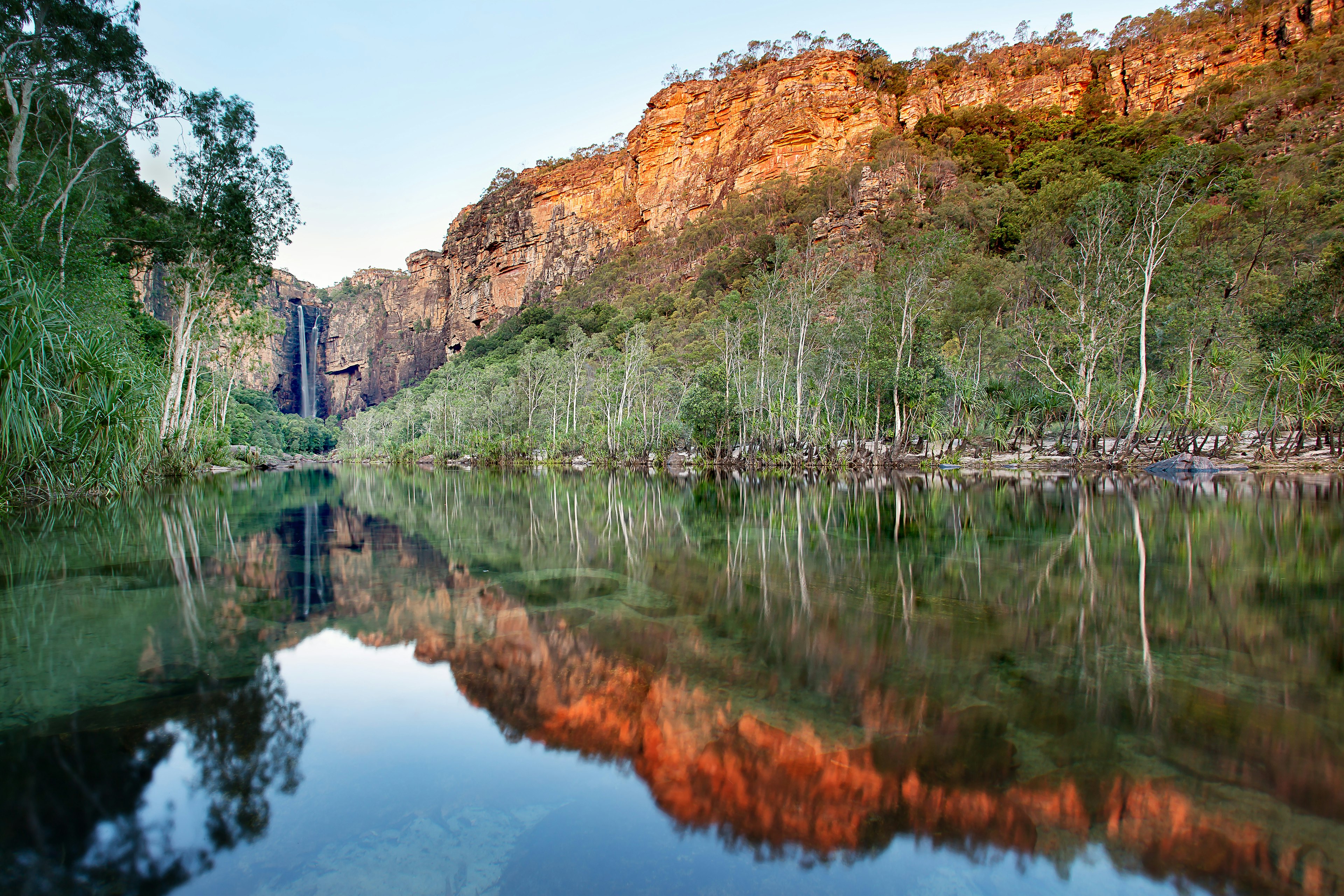 Glassy water is flanked by high trees and red cliffs on either side, with a waterfall at the furthest point from the camera. Kakadu National Park, Australia.