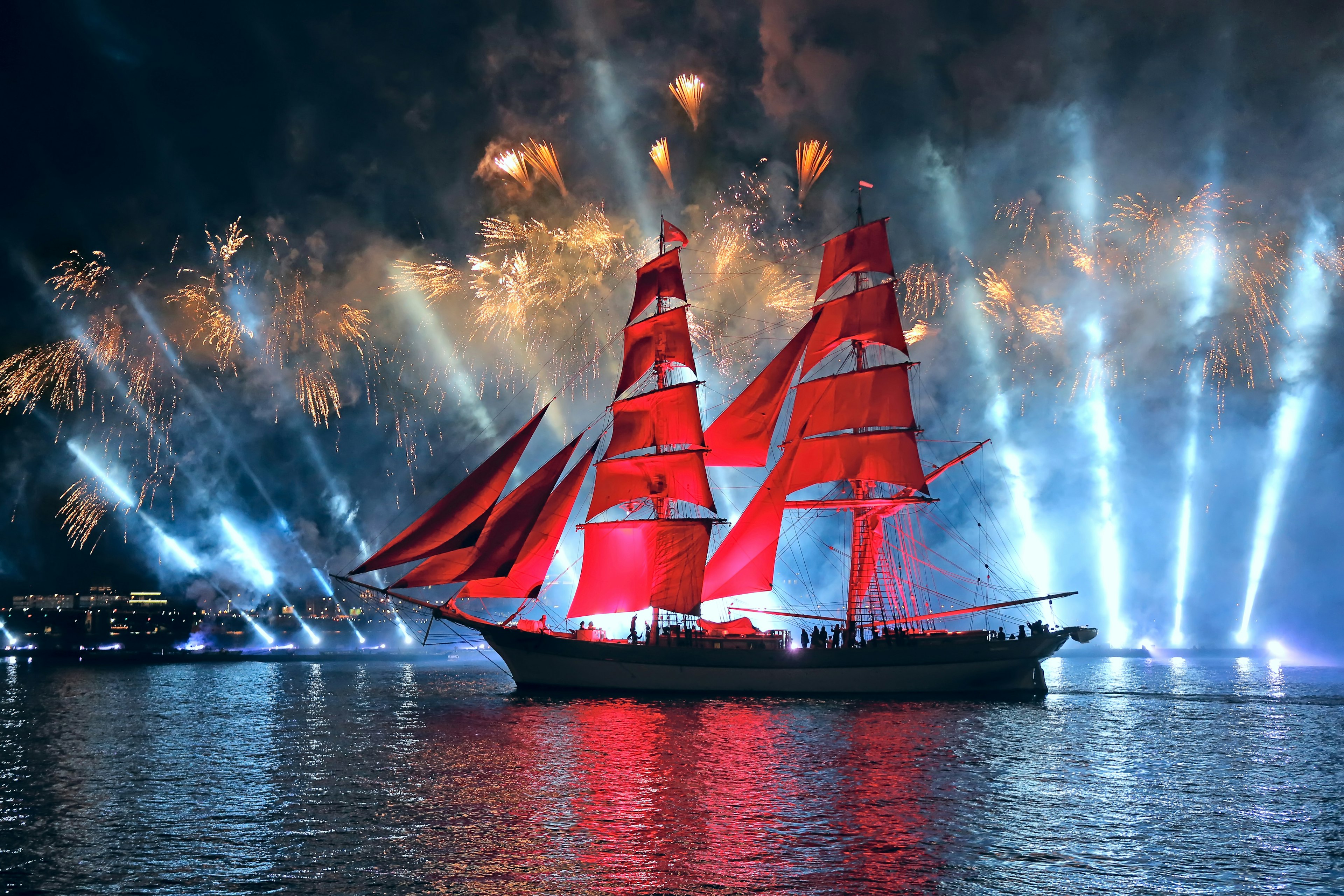 A sail boat on a river at night. It has large red sails and is backlit by bright lights