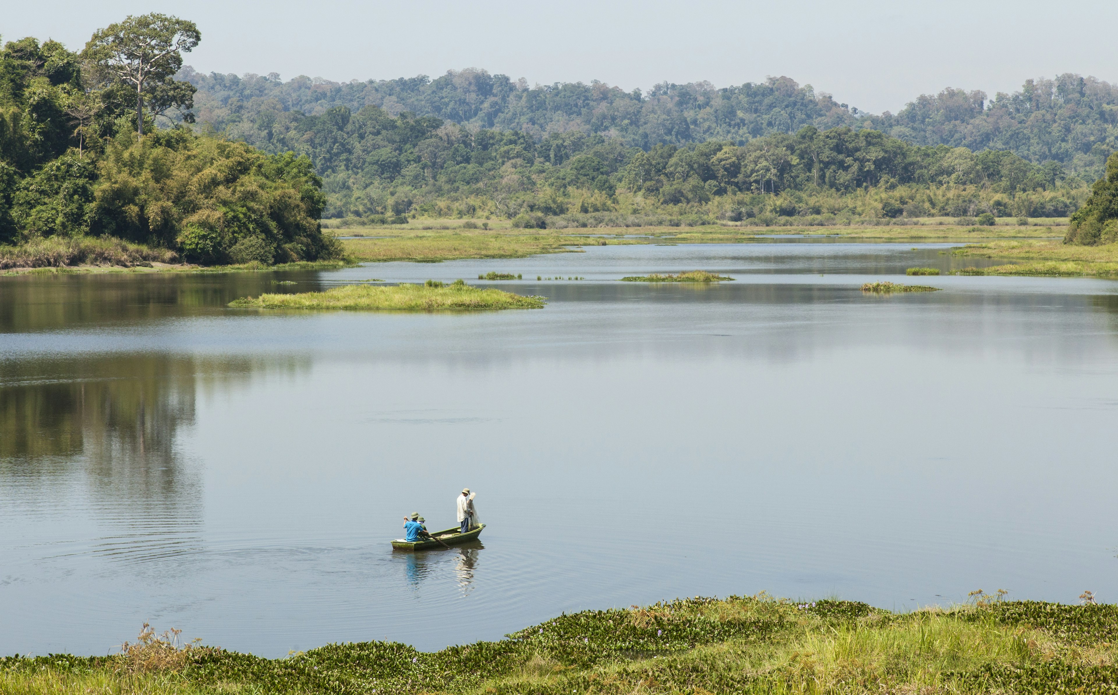 A small wooden boat containing two fishermen floats on a vast lake surrounded by green mountains in Cat Tien National Park, Vietnam.