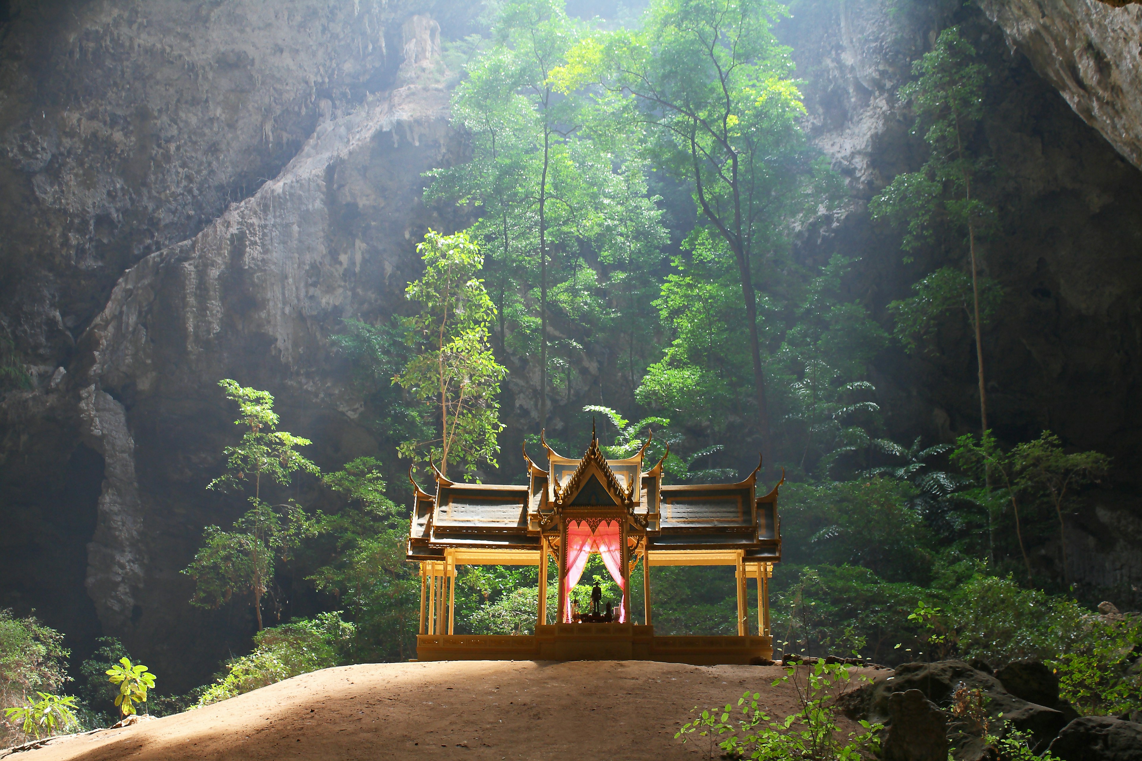 Phraya Nakhon Cave is a tourist attraction In Sam Roi Yot National Park at Prachuap Khiri Khan province in Thailand. A temple stands in the middle of a cave surrounded by trees, with light shining down from above.