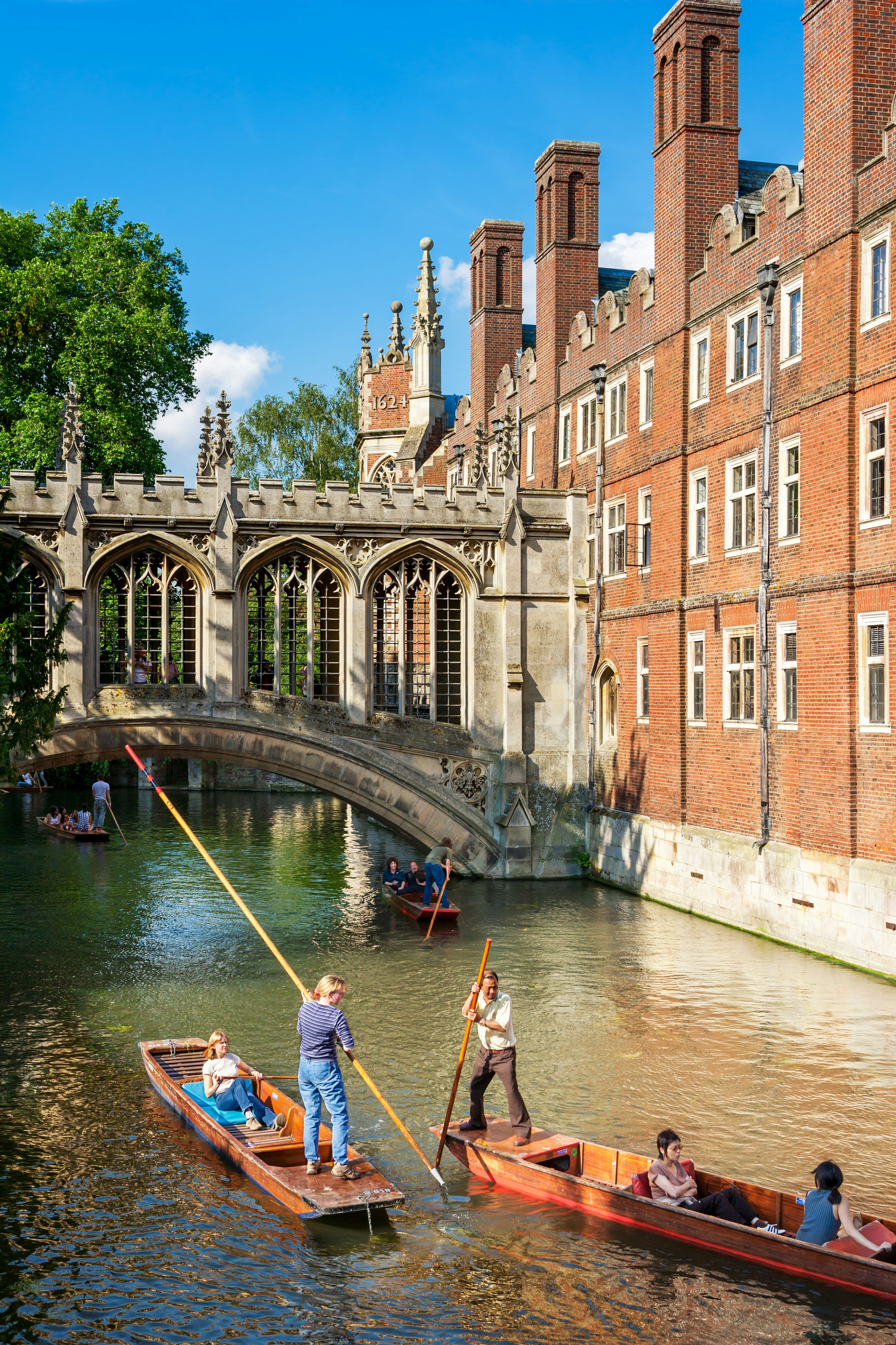 Flat-bottomed boats being pushed along by large wooden poles float down a river under an ornate stone bridge