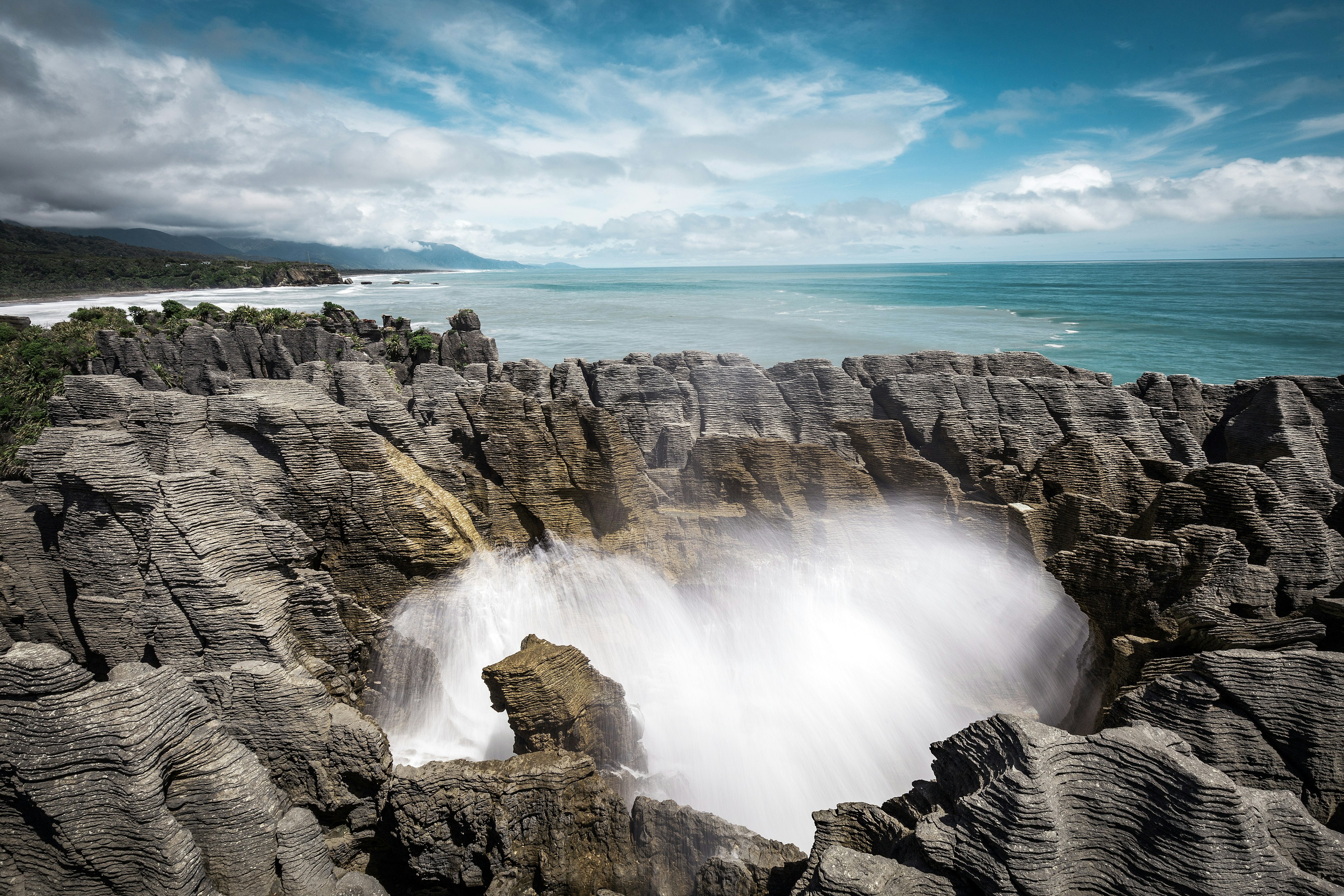 Formations of grey rock, seemingly made of thin sheets of horizontal 'pancakes' rise up into the sky; in the middle of the formation is a blow hole and water is shooting up. In the back ground is the Tasman Sea.