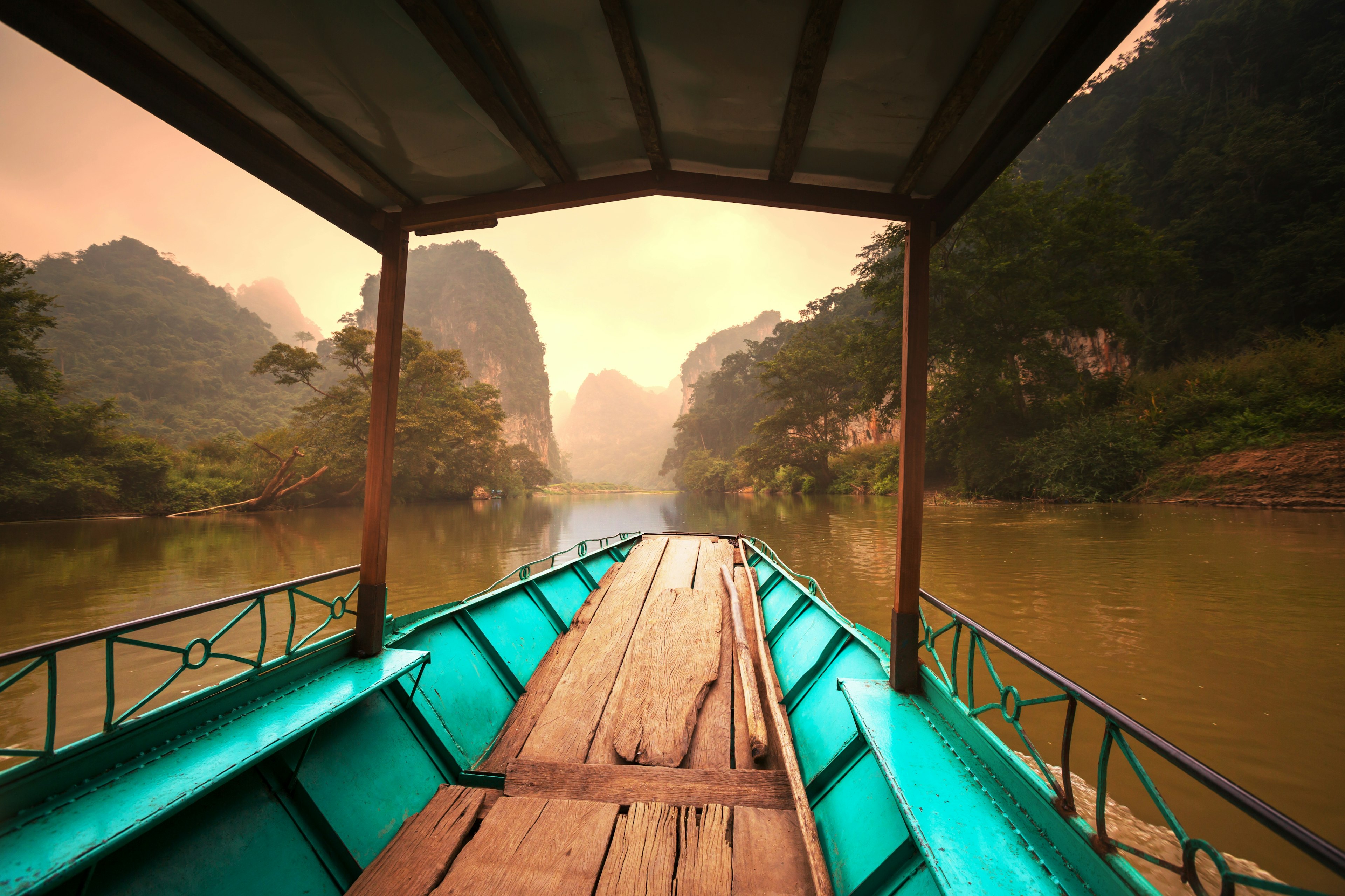 An empty boat floats along in Ba Be National Park. Beyond the boat the shimmering still waters of the lake is visible, which is backed by numerous mountains, most of which are covered in forest.