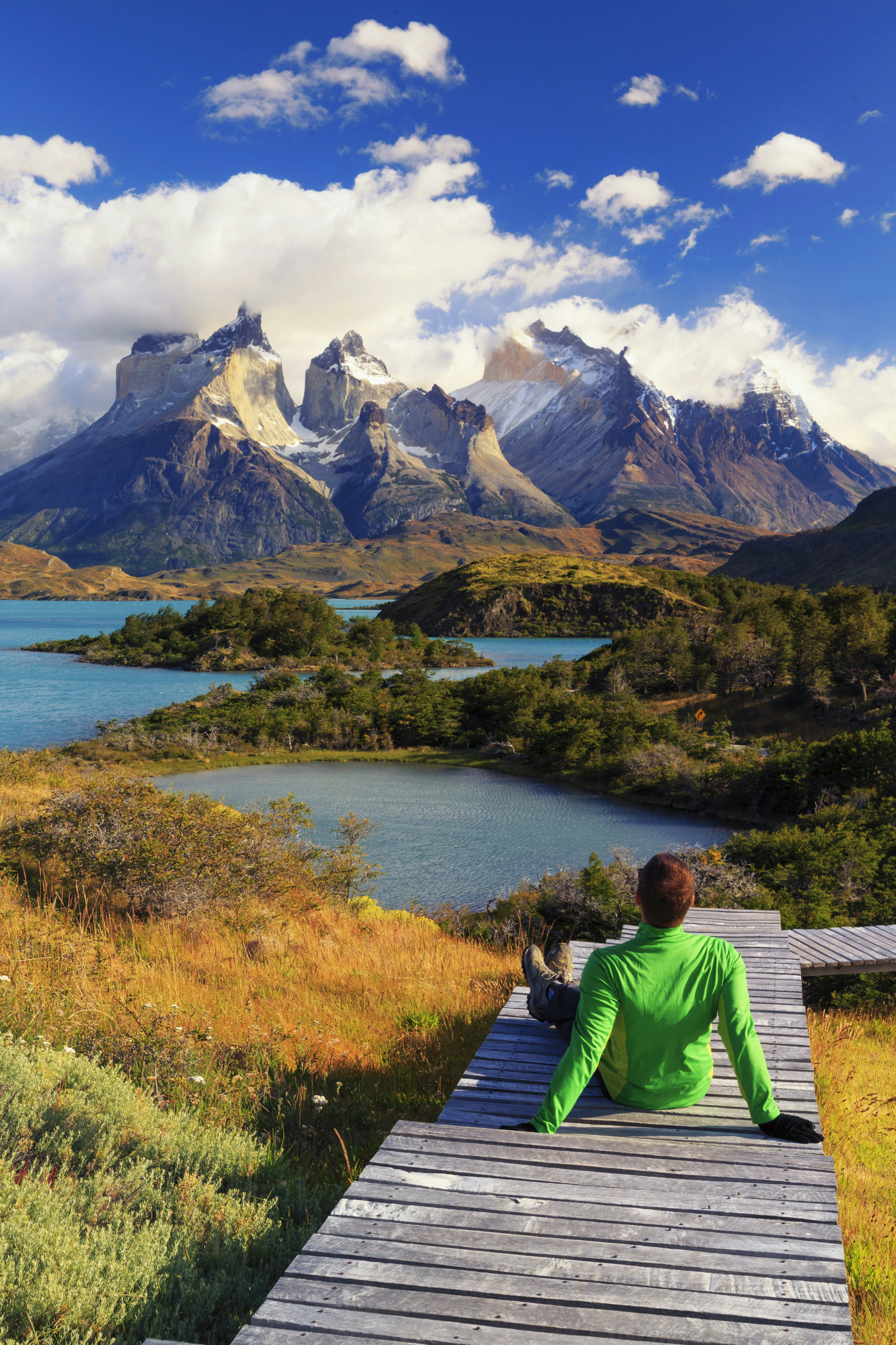 A hiker sits on a boardwalk above a lake surrounded by mountain peaks - Torres del Paine National Park