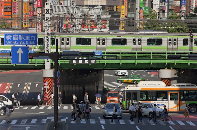 A train travels over a bridge above roads with pedestrians, cars and a bus below