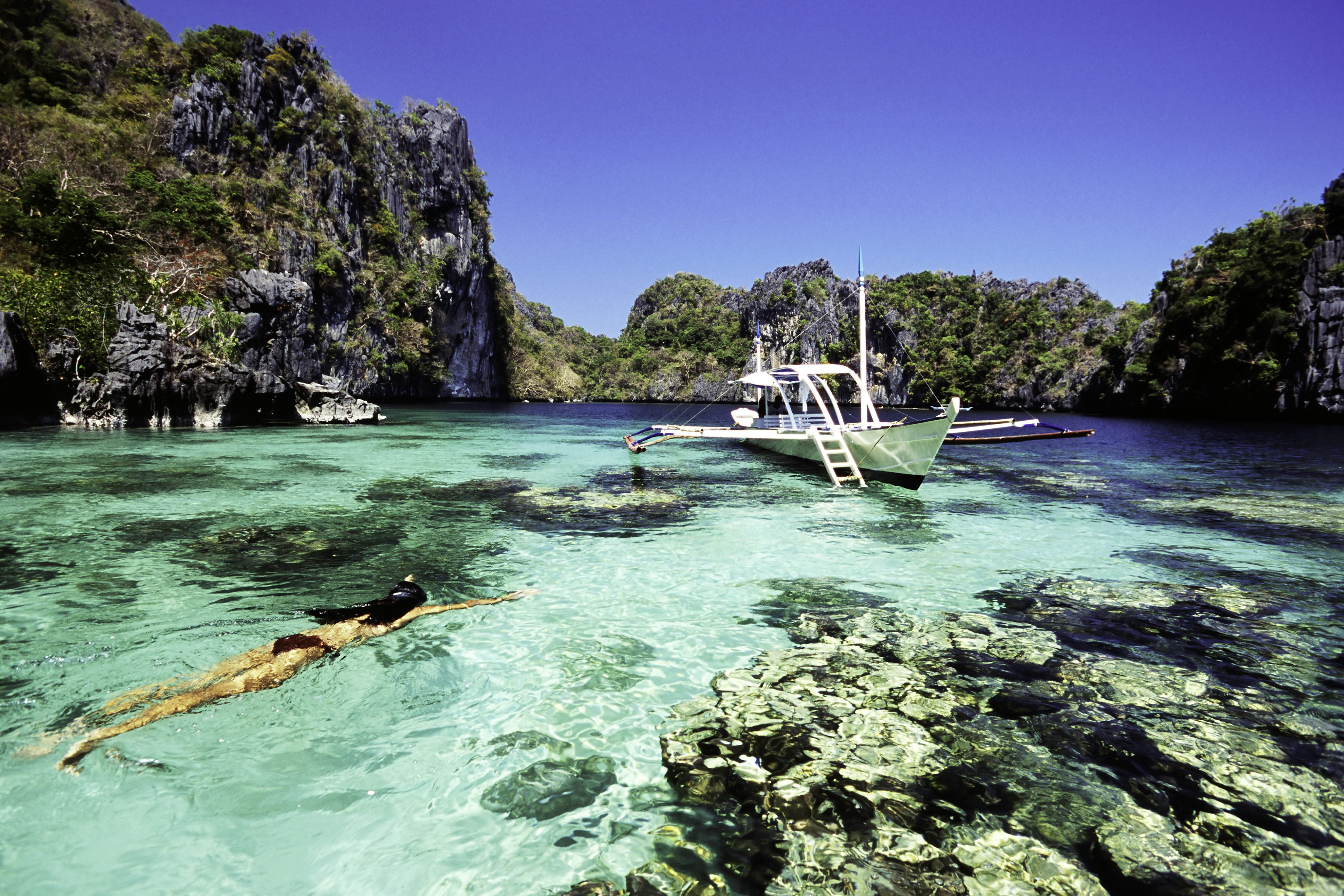 A woman snorkels in clear water next to a boat with pontoons in a rocky lagoon