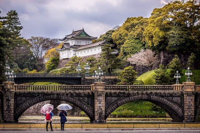 Two women stood beneath umbrellas stand in front of a bridge at the Imperial Palace in Tokyo