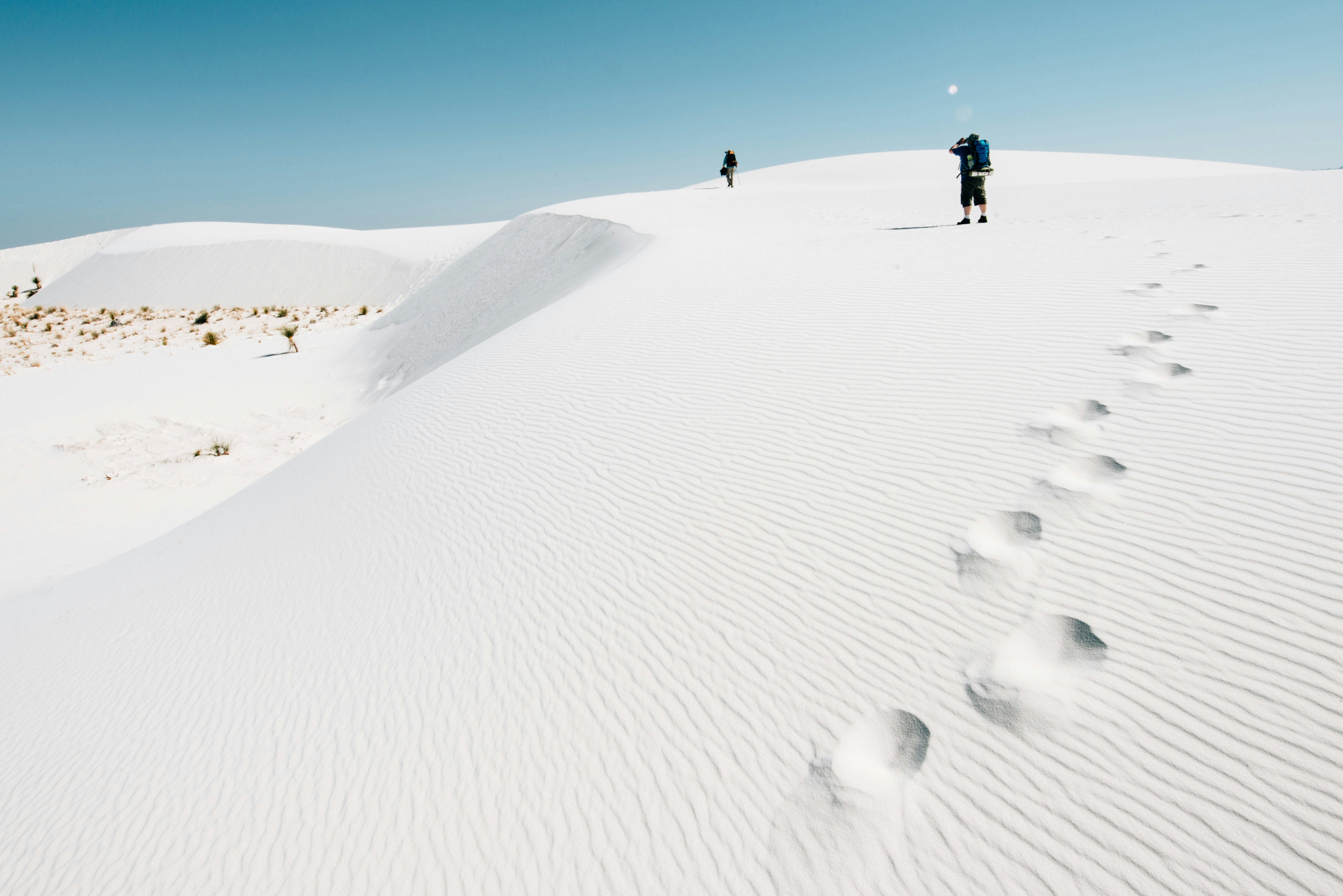 People in backpacks are seen in the distance on wind-blown dunes of white sand. Footprints are visible on the dunes.