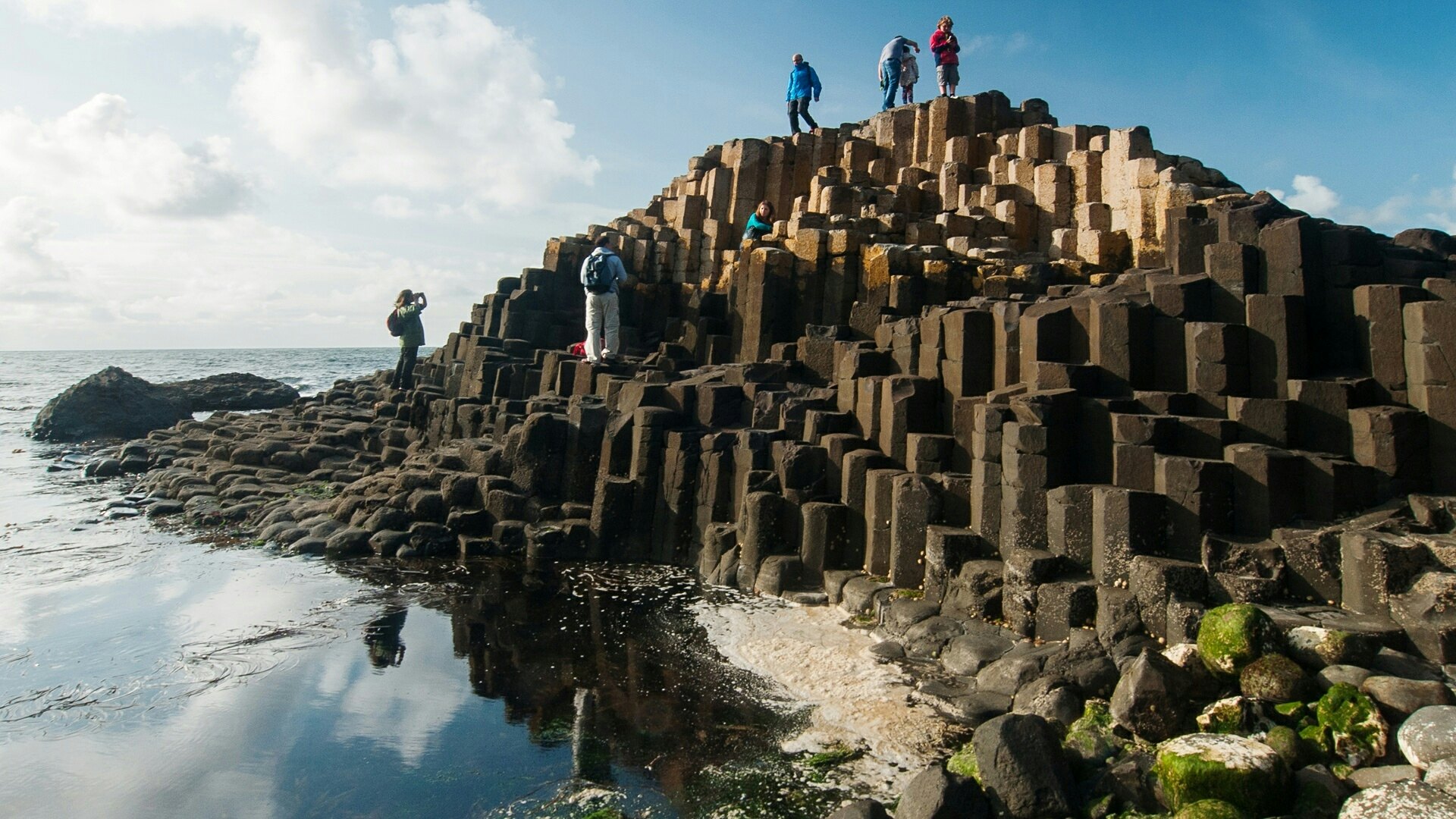 People walk on a seaside rock formation of hexagonal basalt columns.