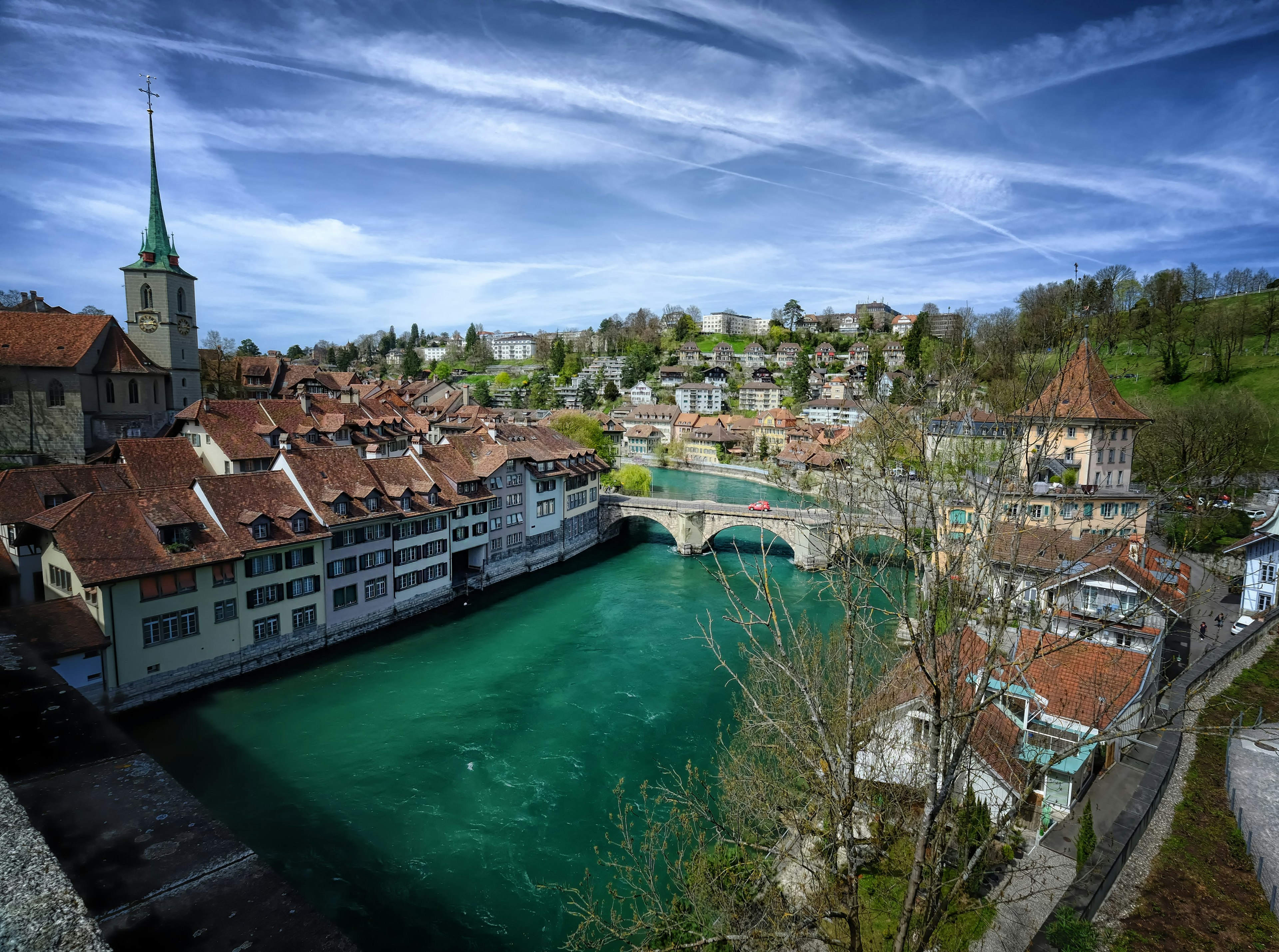 A blue green river flows though a historic city. It flows under a stone bridge with three arches and on each side of the river there are medieval-style buildings. On the left is a church with a tall, green spire.