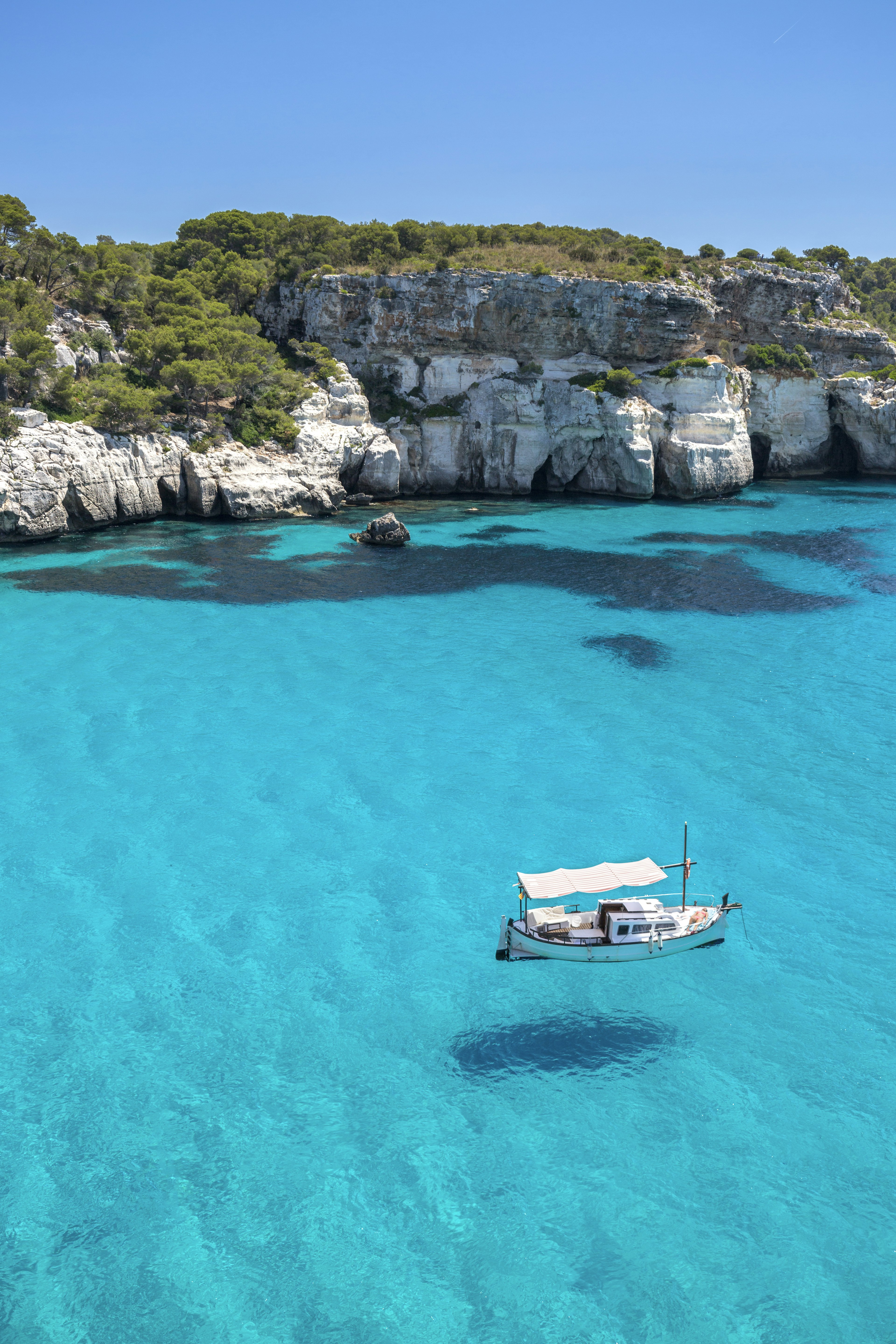 A small sail boat sits in pristine clear turquoise waters near a rocky coastline