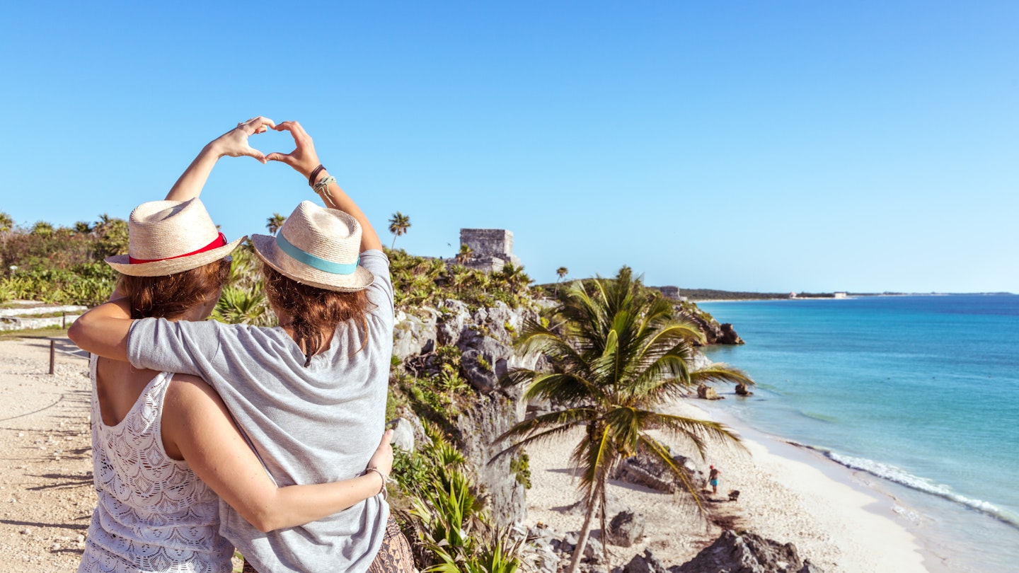 two women with their arms around each other create a heart with their hands while looking at a Mayan ruin on the beach