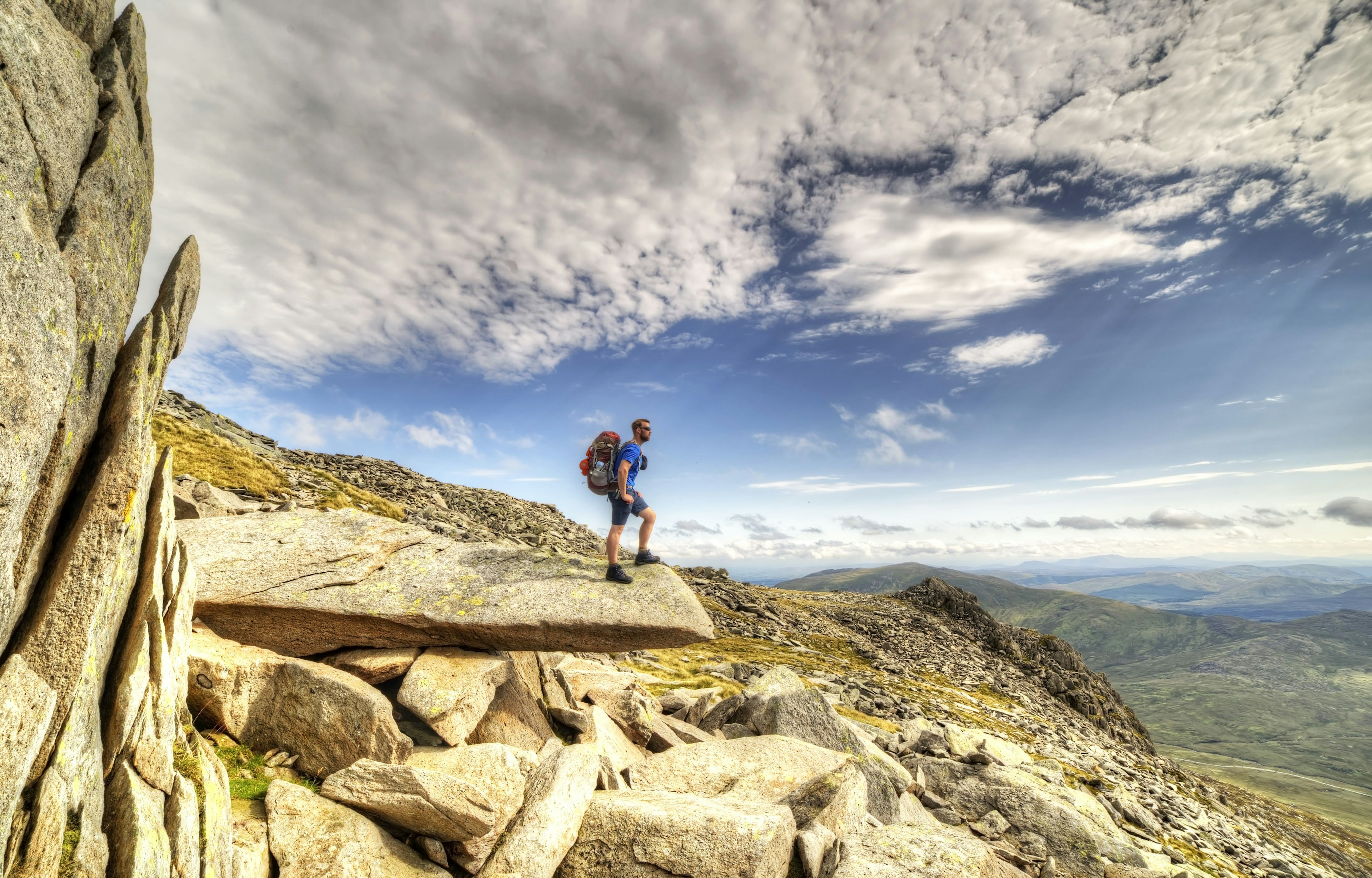 A man in hiking gear carrying a backpack stands on a rocky outcrop with rolling hills in the distance