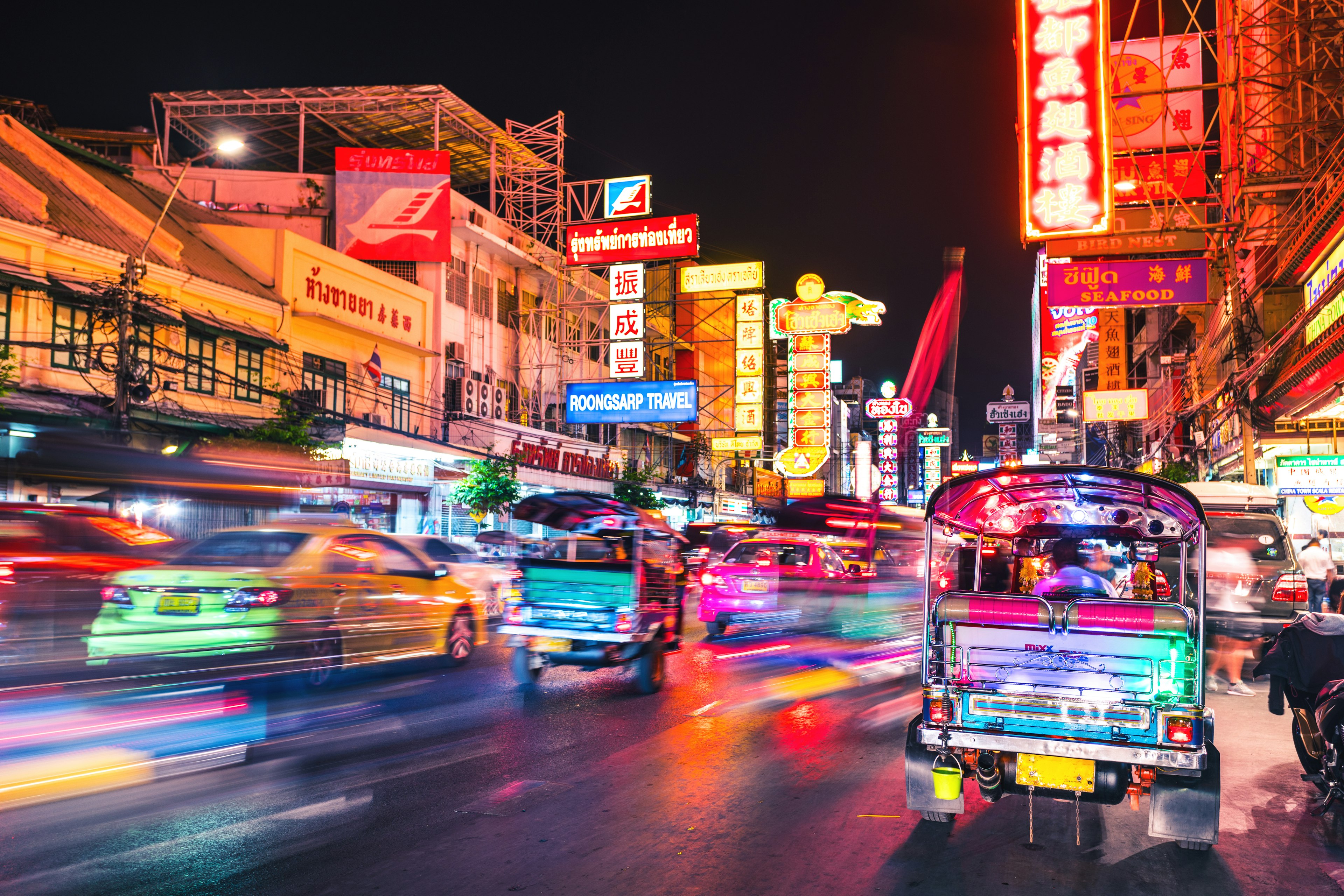 A glaze of neon as tuk-tuks and other traffic streams down a lit-up street at night.