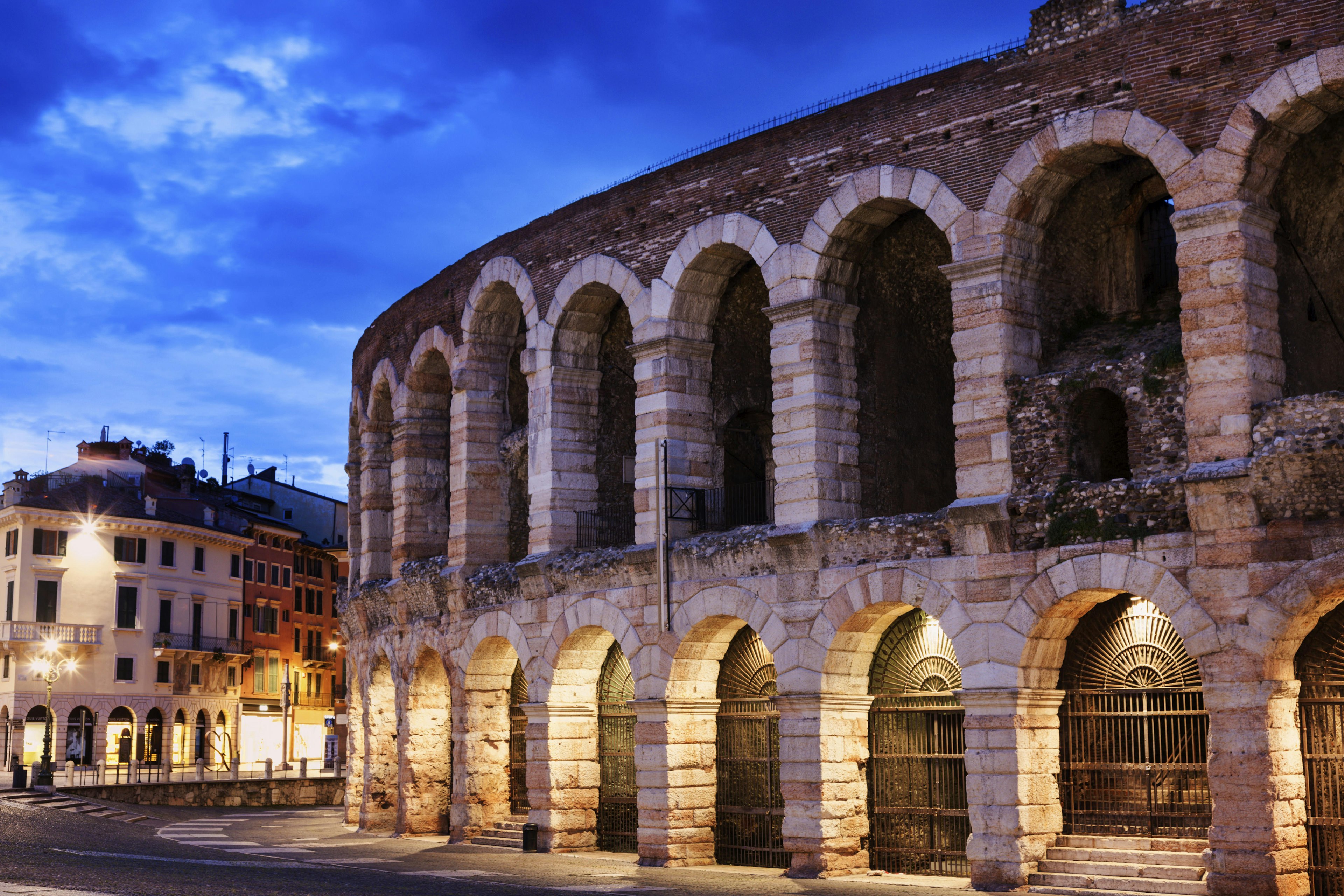 The stone exterior of a large arena at dusk, with low lighting under each of the archways