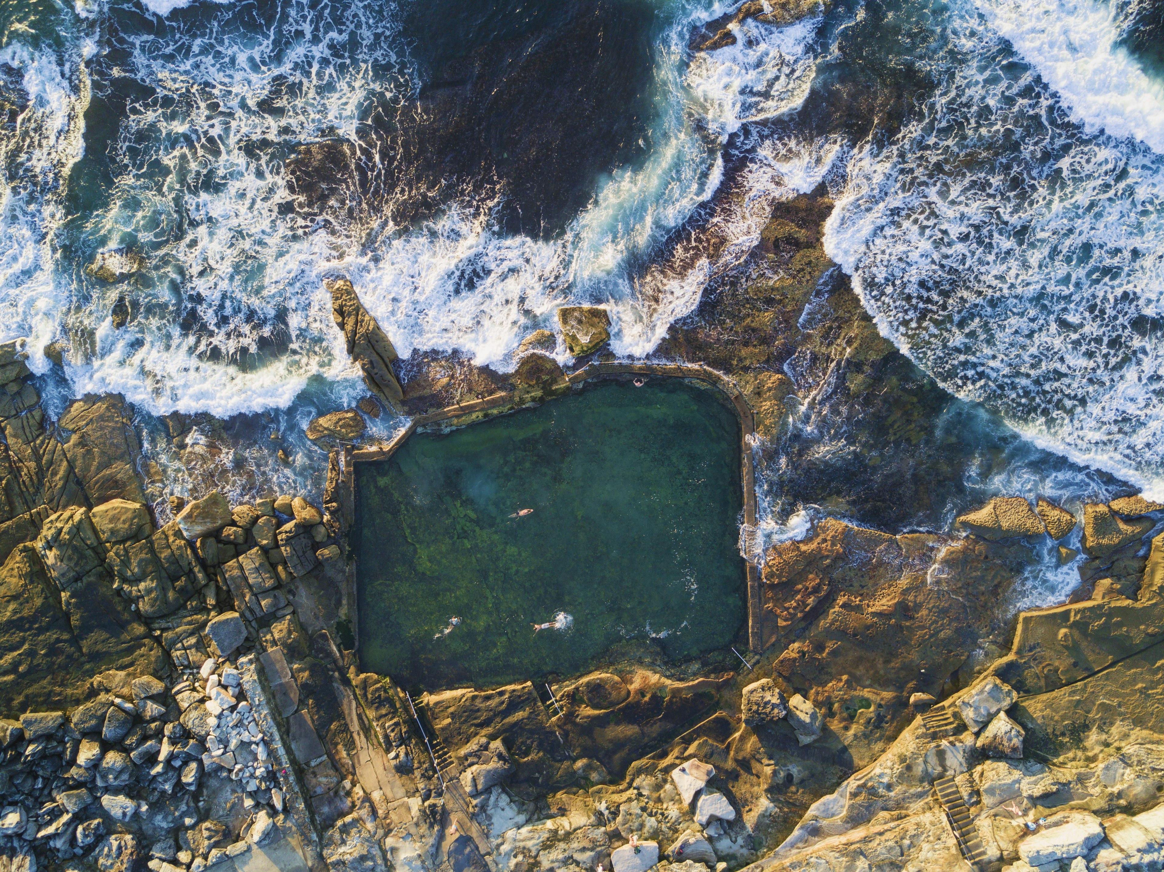 Overhead shot of people swimming as waves crash into the Mahon rock pool on a sunny day