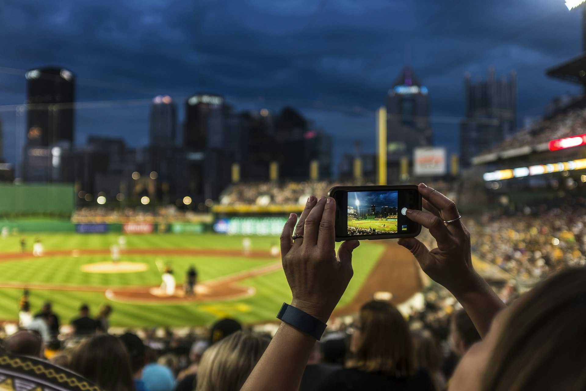 A woman holds a phone to photograph a Pirates baseball game at PNC Park, in front of the Pittsburgh skyline at night