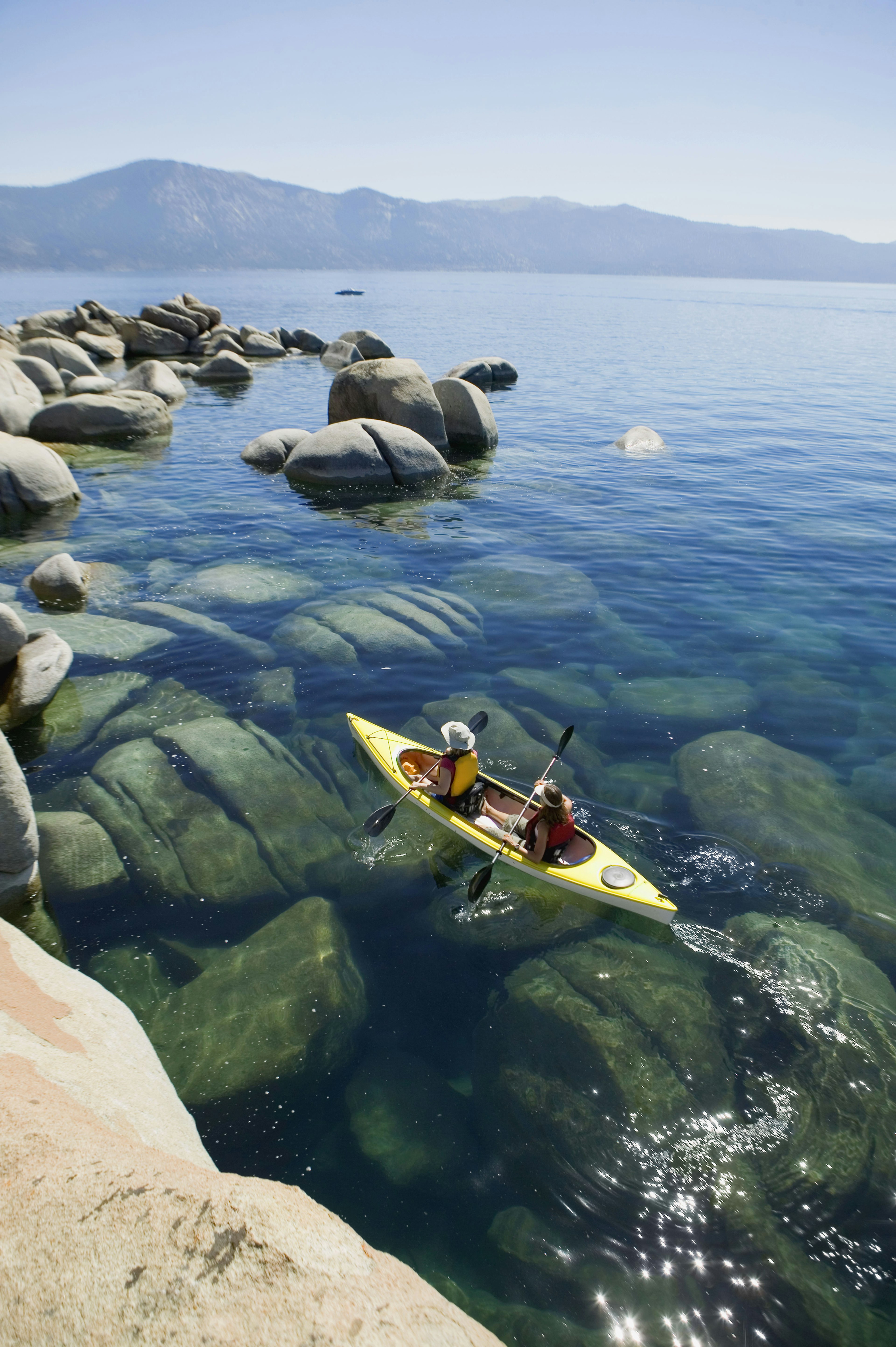 Two people row in a small yellow kayak near the shore of a vast lake on a sunny day