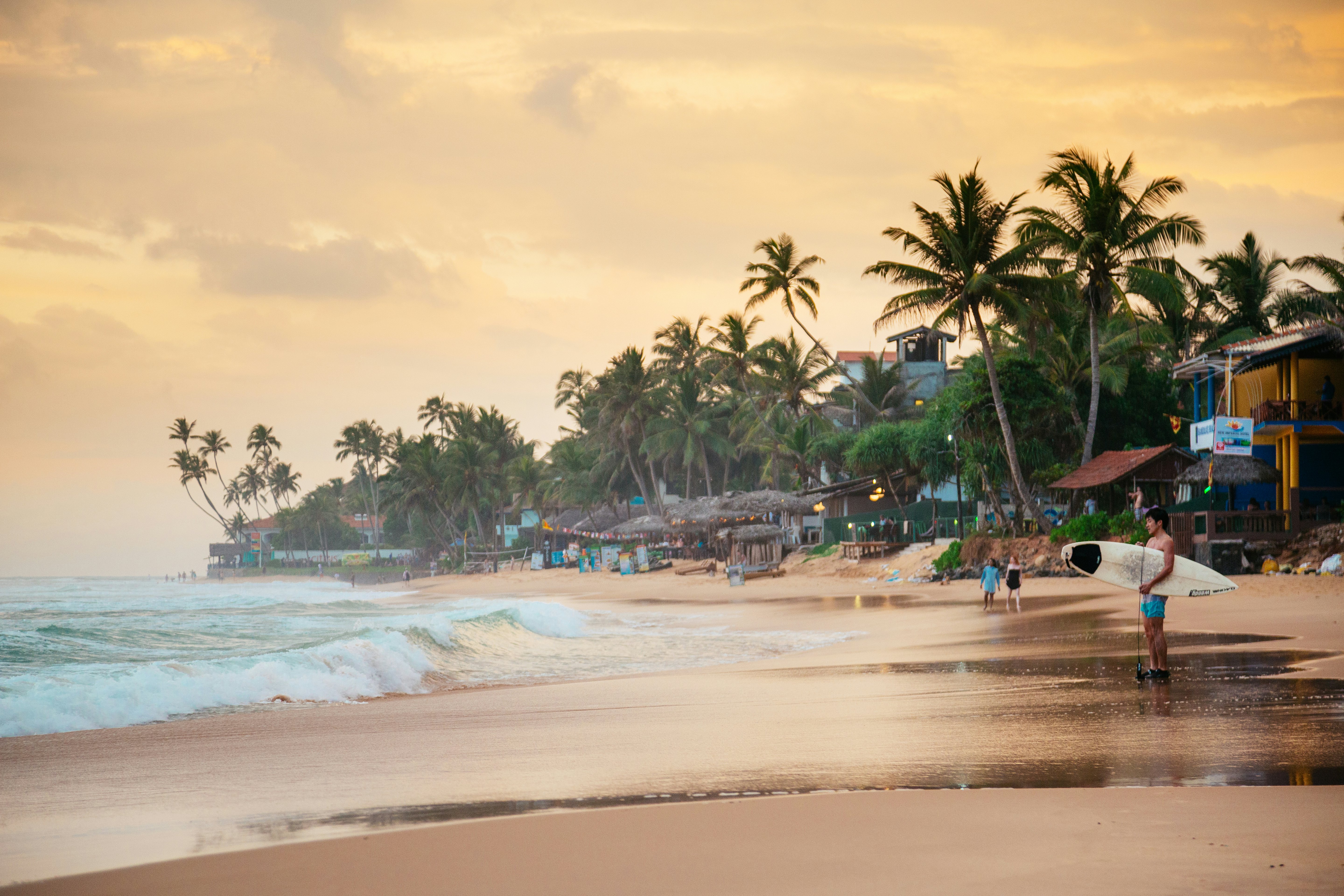 A surfer stands on the sand at Narigama beach in Hikkaduwa during sunset.