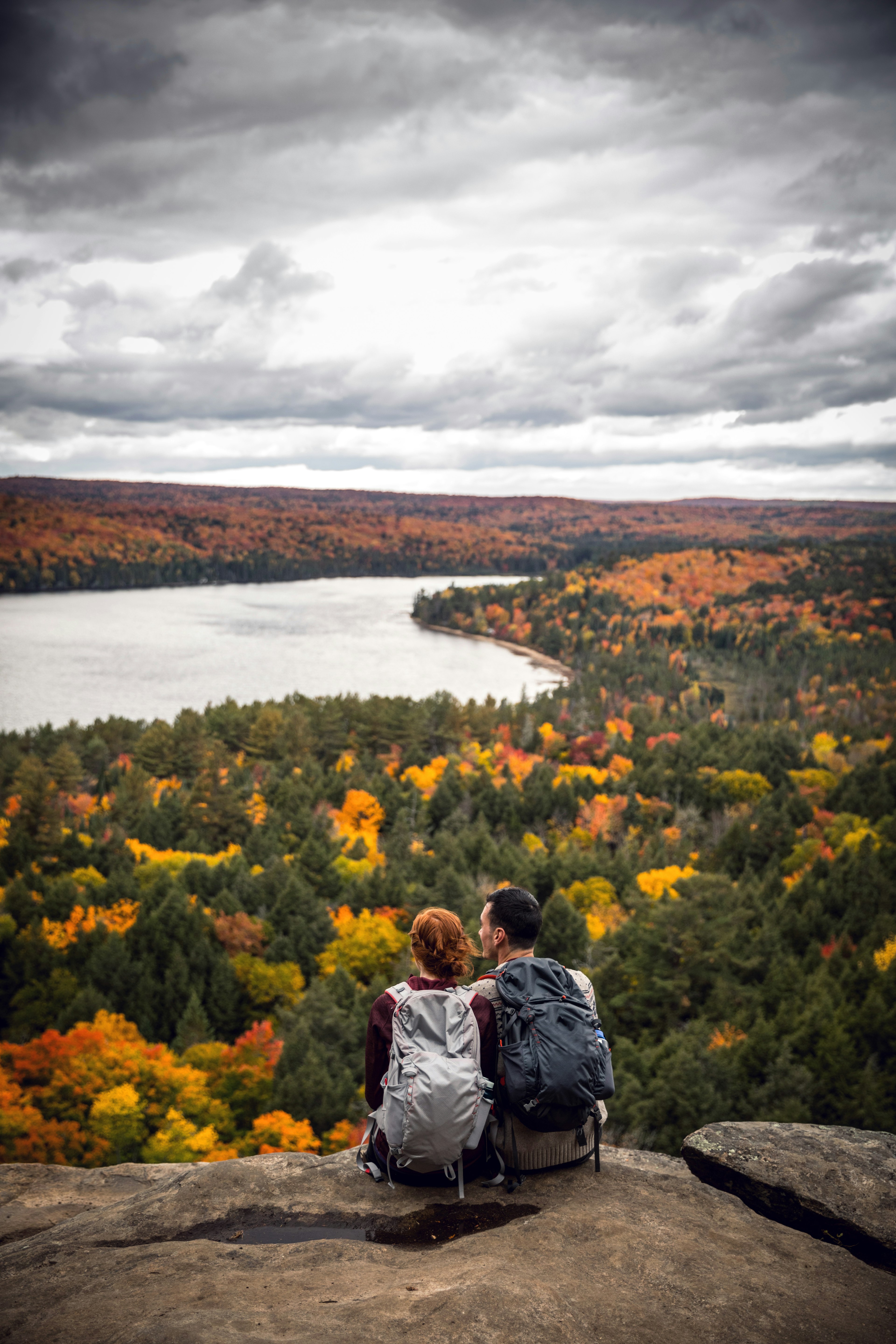 Couple sitting on a rock, overlooking a forest of fall colors and a lake in Algonquin Provincial Park, Canada