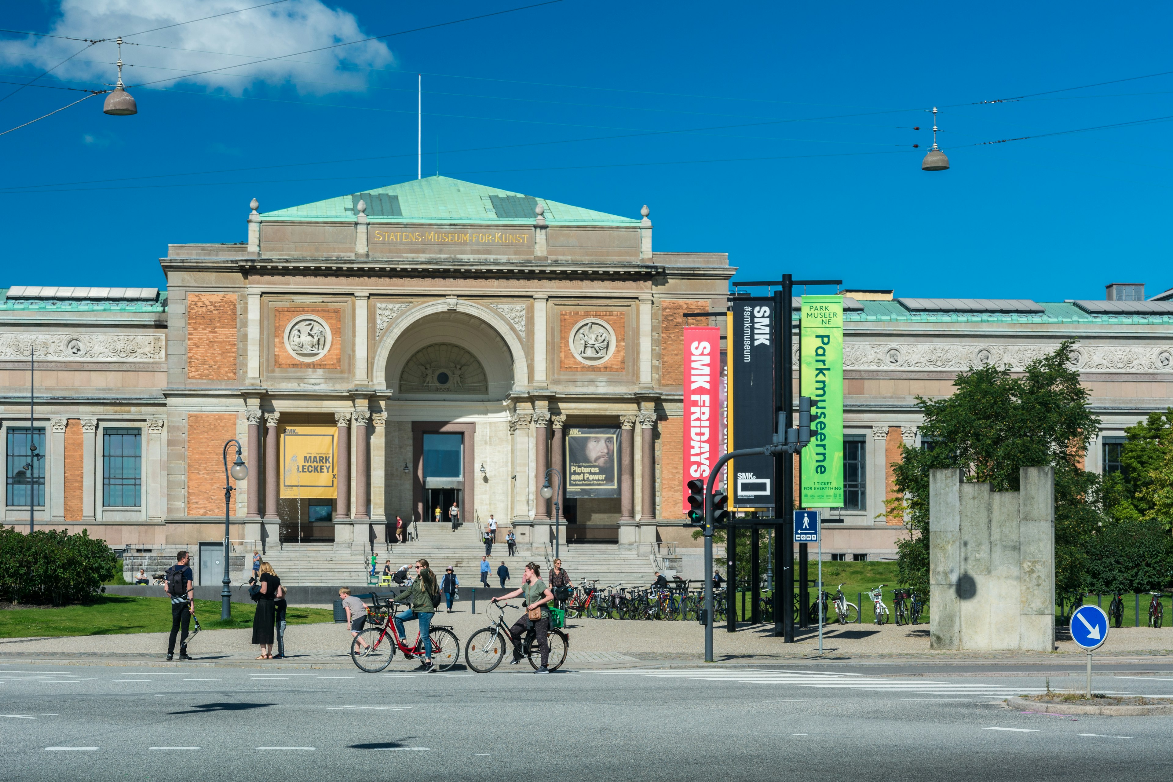 People walk and bike in front of a large, historic-looking building with a copper roof