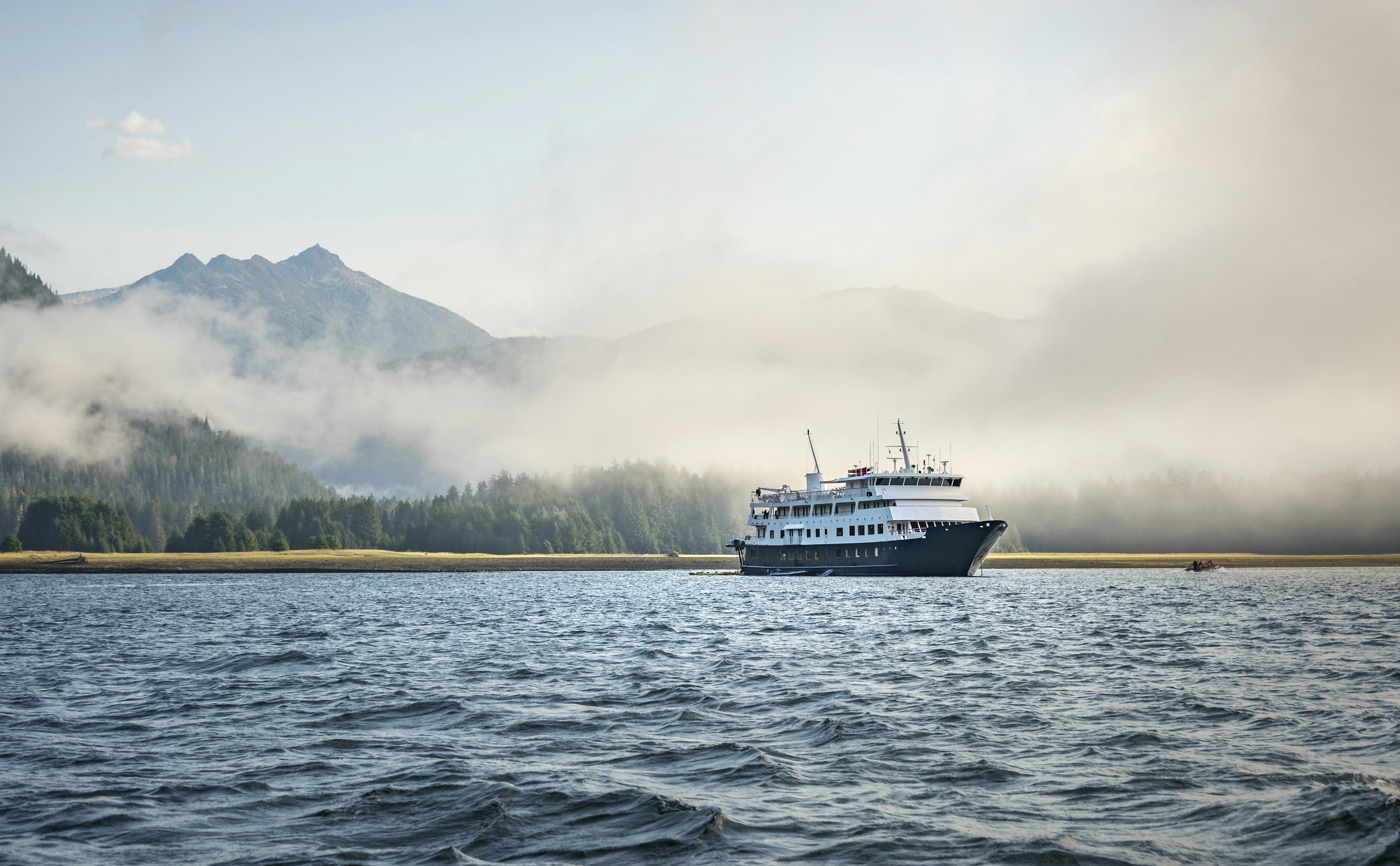 A passenger ferry sails on dark-blue waters, with a heavily forested, mountainous landscape blanketed in cloud in the background