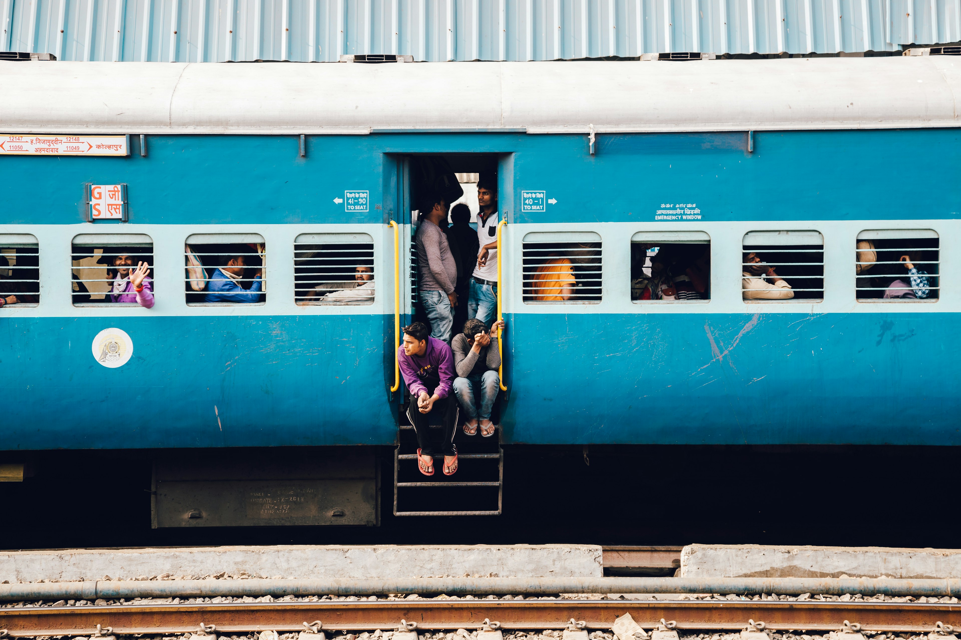 A stationary blue train in India, with passengers sitting and leaning out of the doorway and looking through the train's windows.