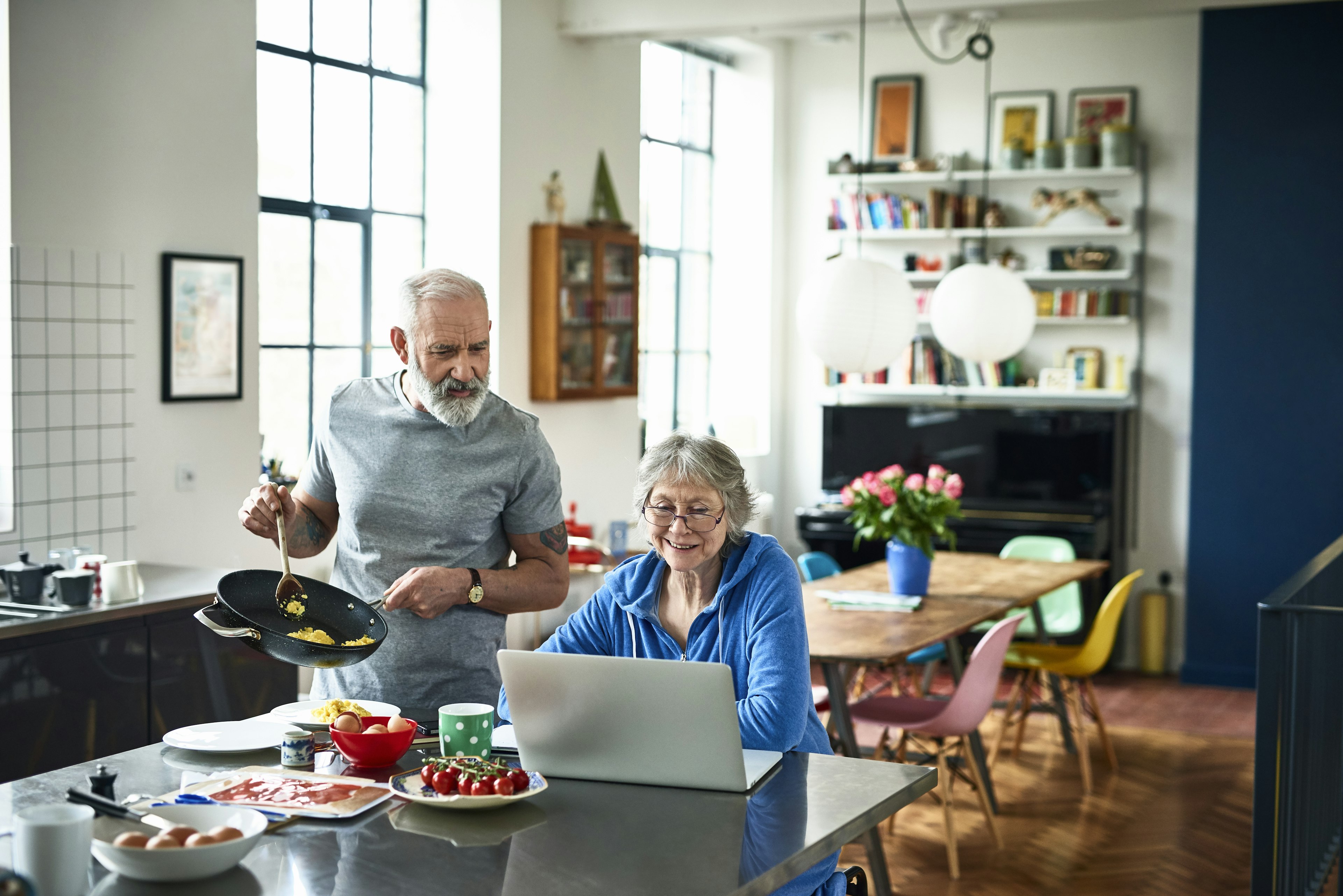 Senior woman using laptop and smiling as man serves breakfast