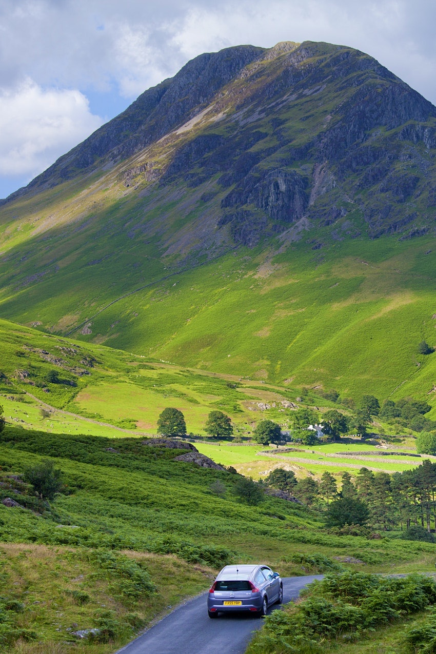 A gray car rides down a single lane road surrounded by green landscape and towards a very tall mountain