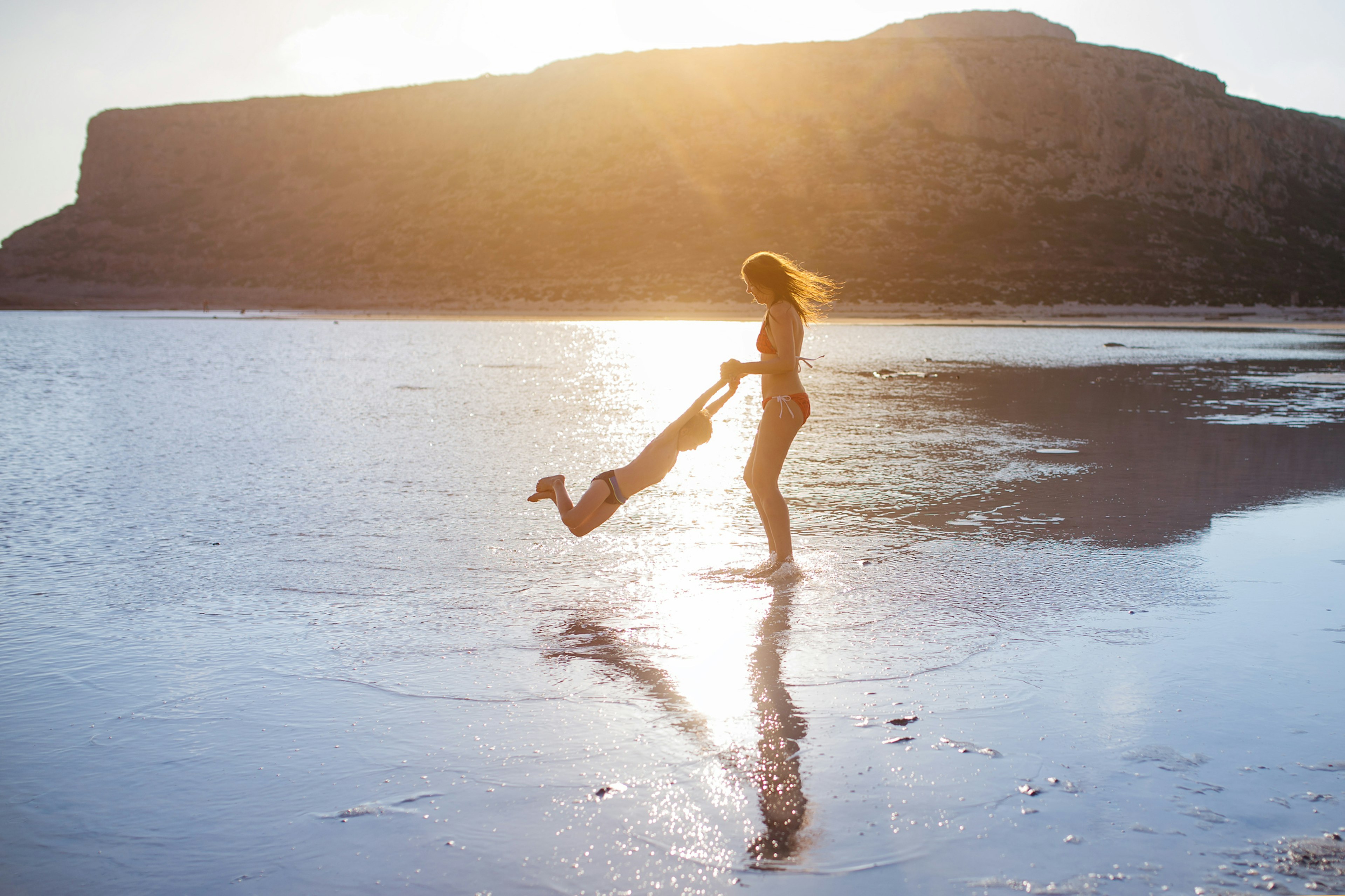 A woman swings a young boy around as they laugh on the beach in Crete.