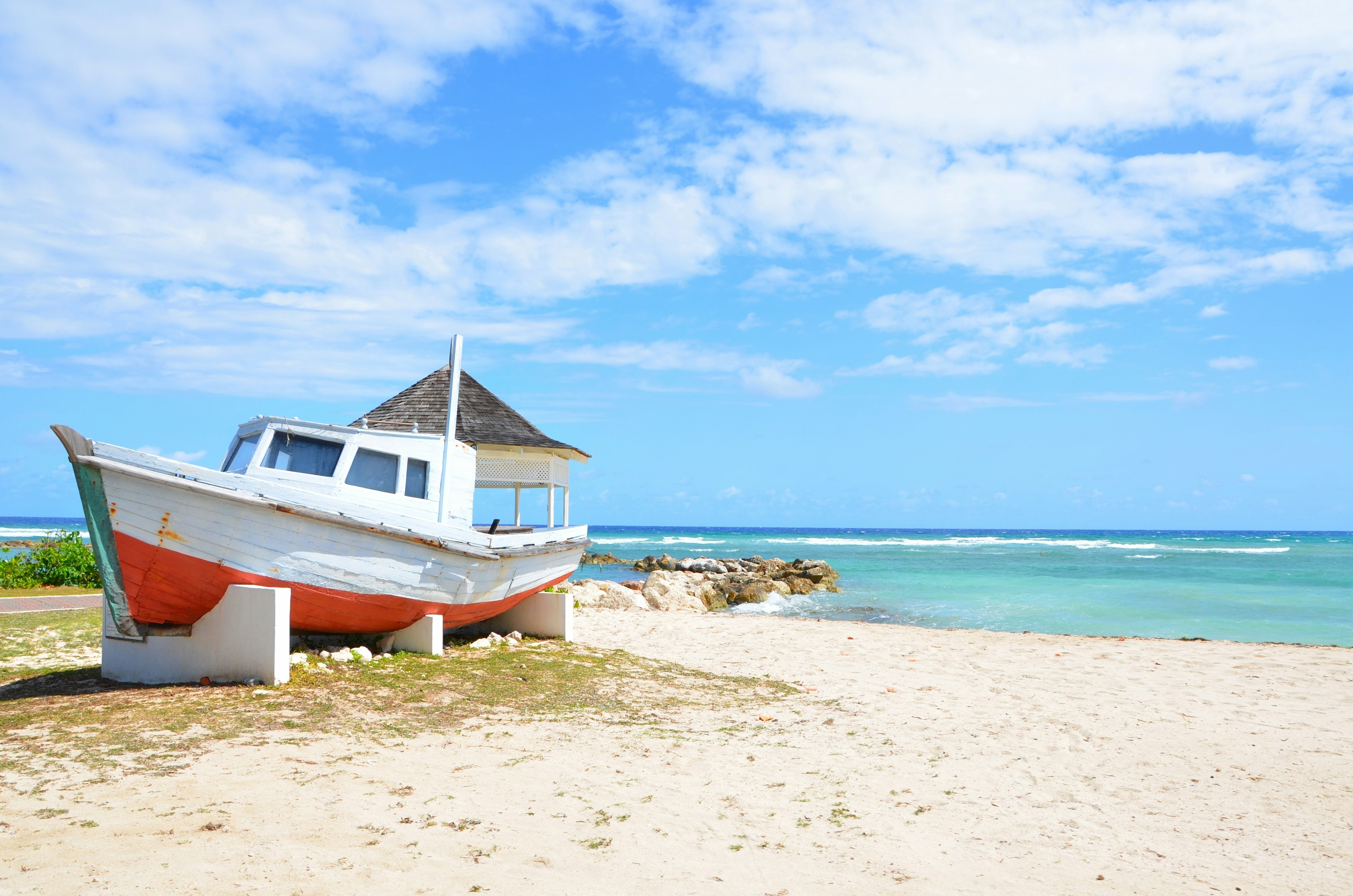 A white boat with an orange bottom is stacked on wooden pillars on the sand. There are rocks jutting into the ocean.