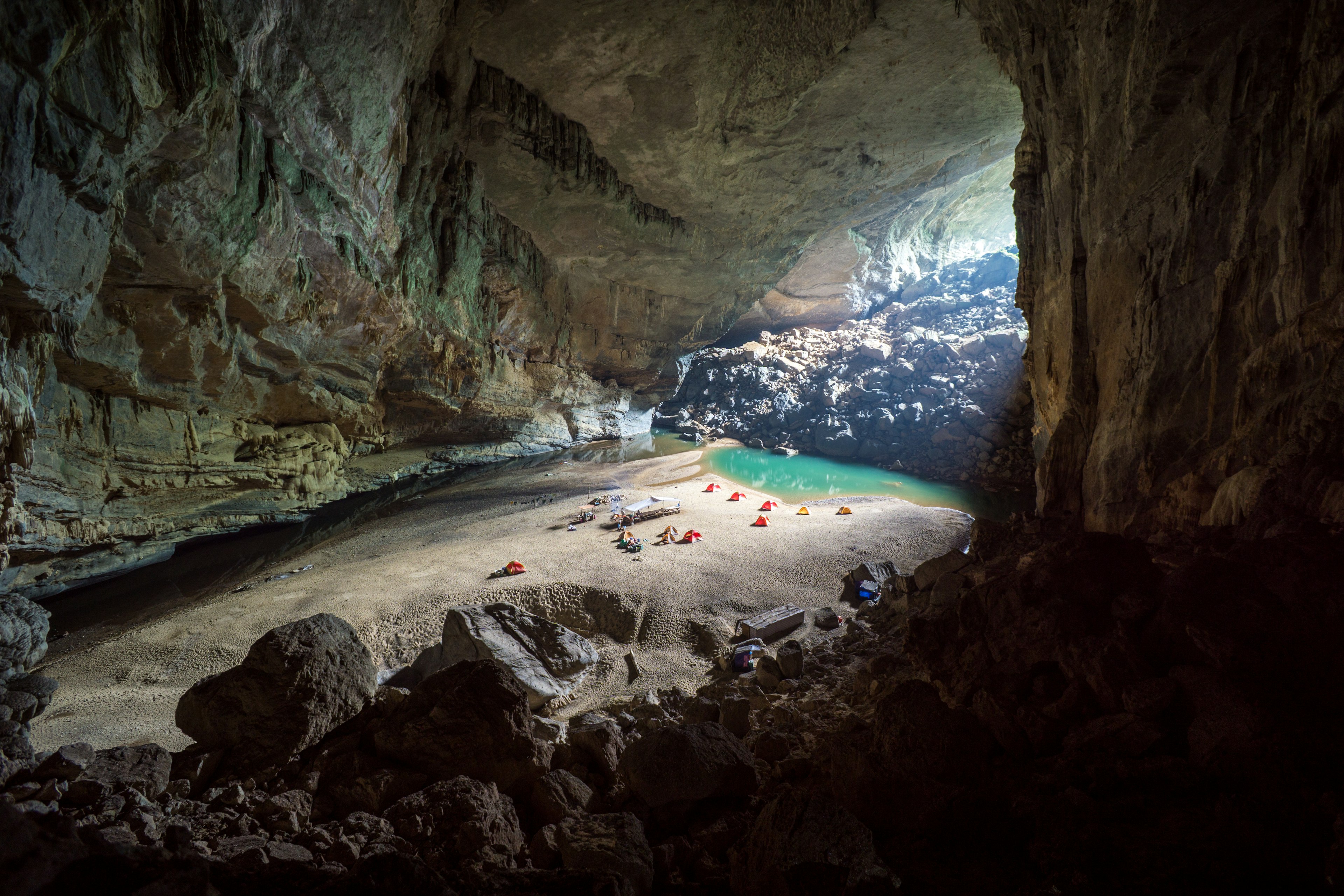 A handful of yellow and red tents can be seen erected on a sand bar near the entrance to a cave