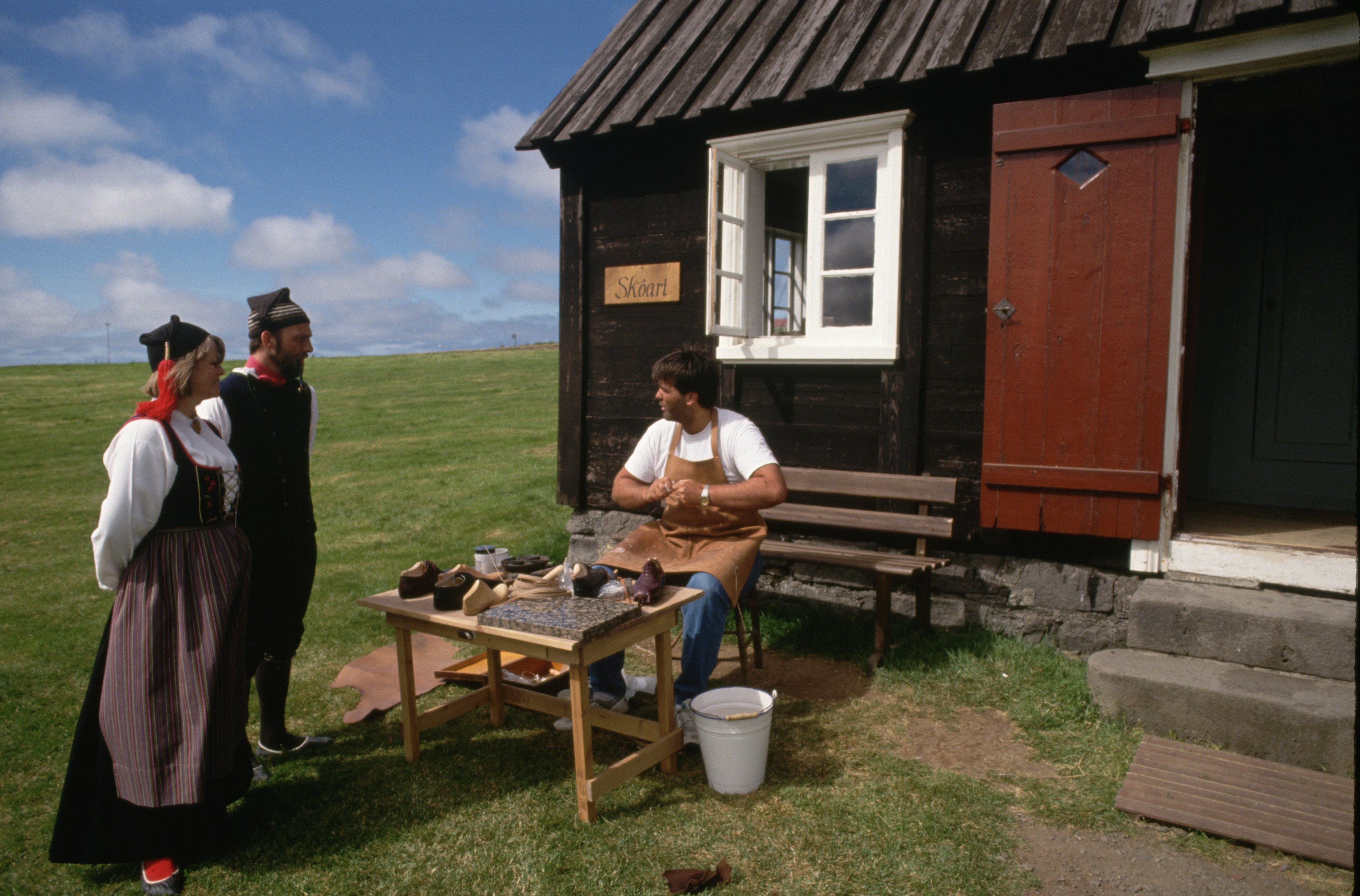 Wearing traditional dress, actors at Arbaejarsafn, or Open Air Museum, talk to a fellow actor at his shoemaking post. Reykjavik, Iceland