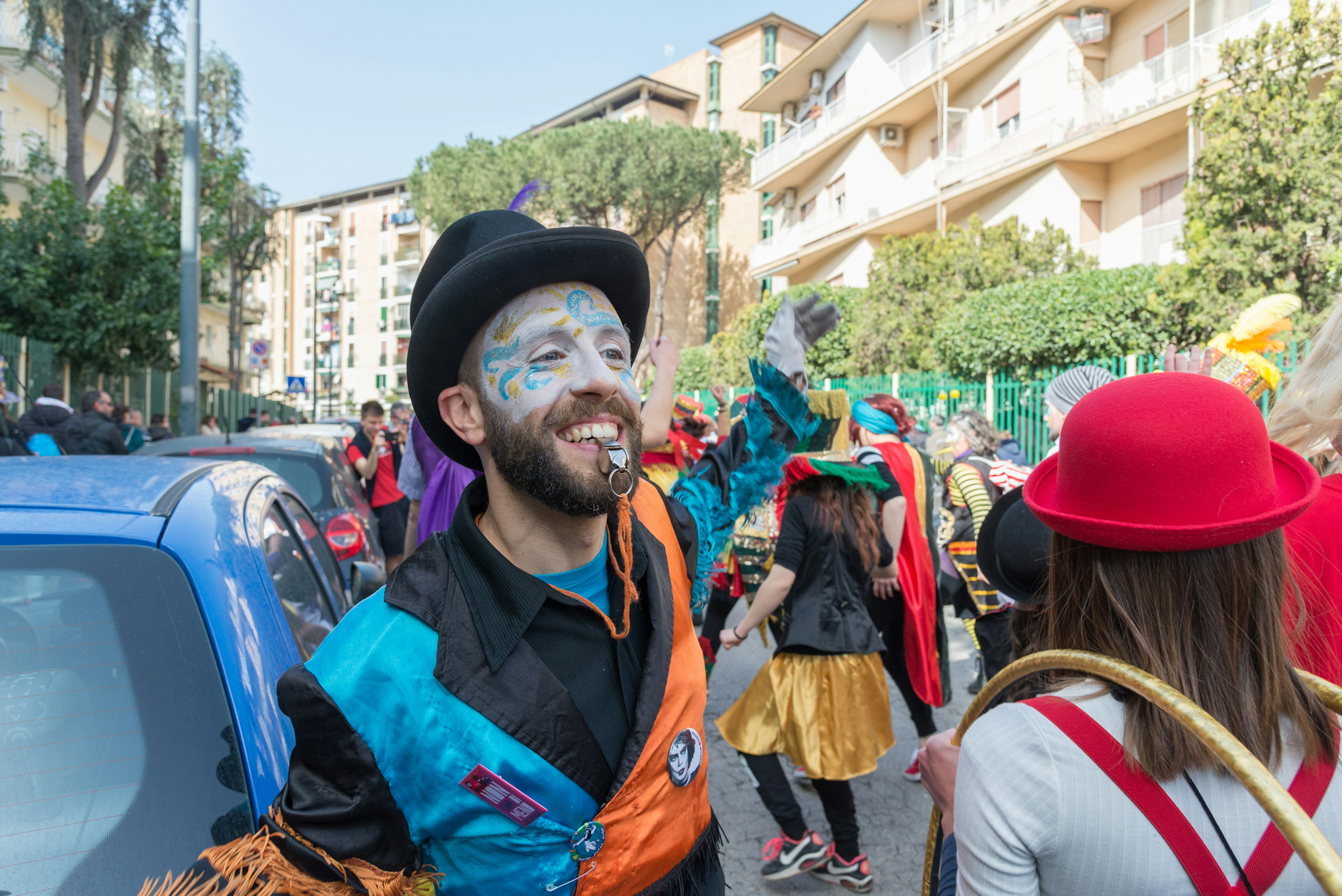 A man covered in colorful face paint smiles as he blows a whistle to direct carnival-goers down a path