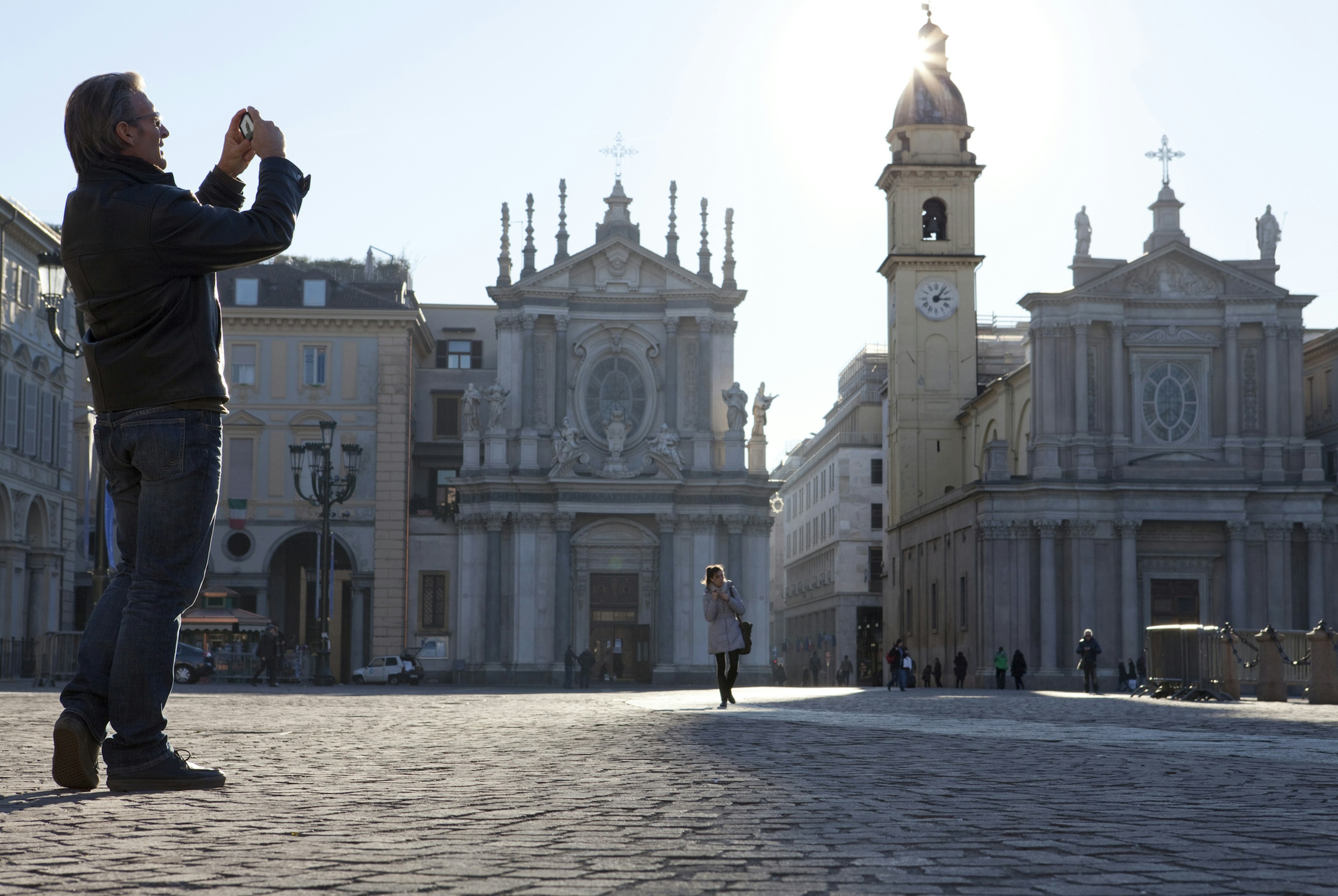 A older man, slicked back hair, glasses and leather jacket, takes a photo on a phone of a cobbled Turin piazza as the sun begins to go down