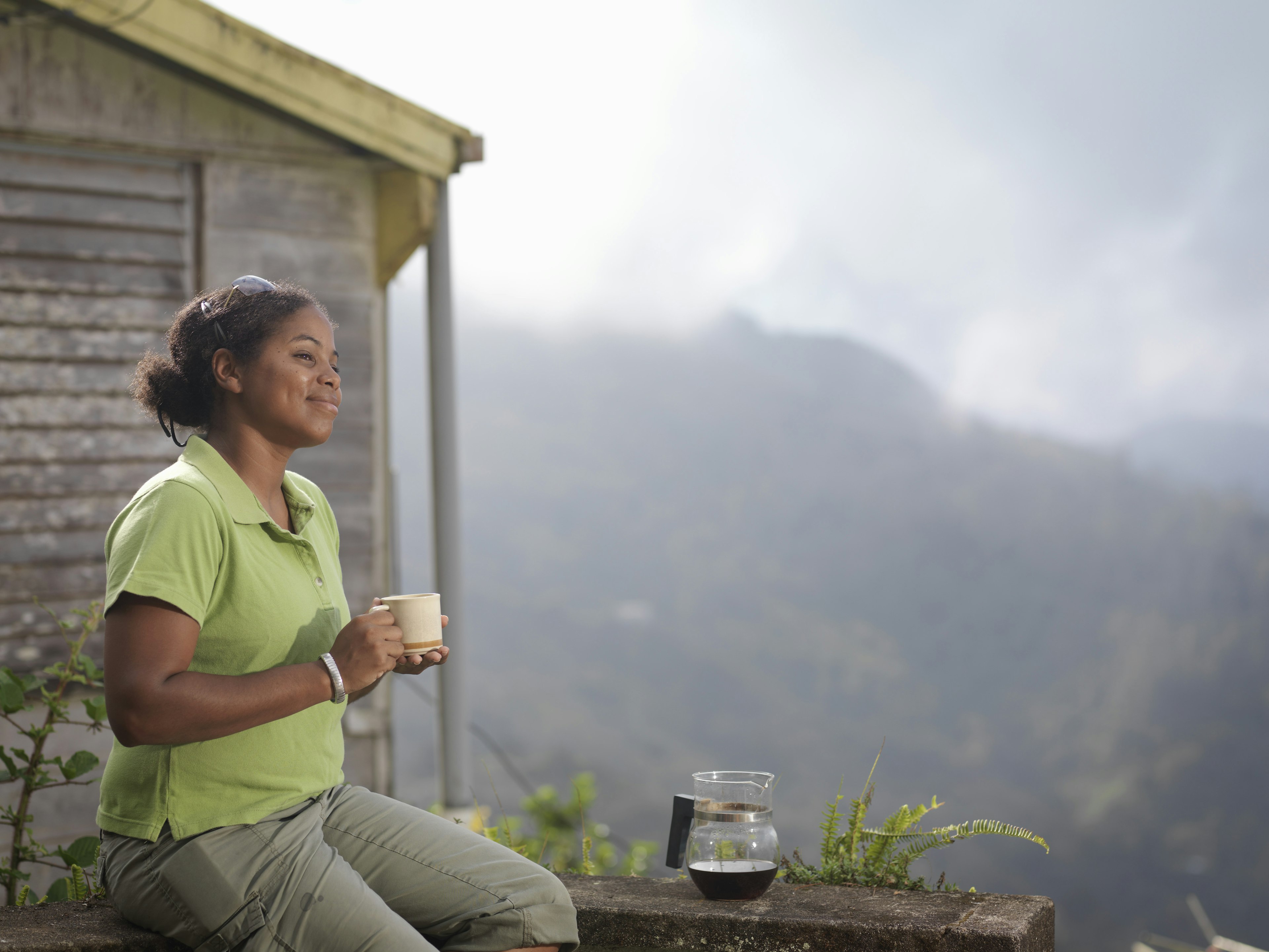 A woman wearing a green top holds a cup of coffee while looking out over the mountains in Jamaica. There is a glass coffee pot on a stone ledge.